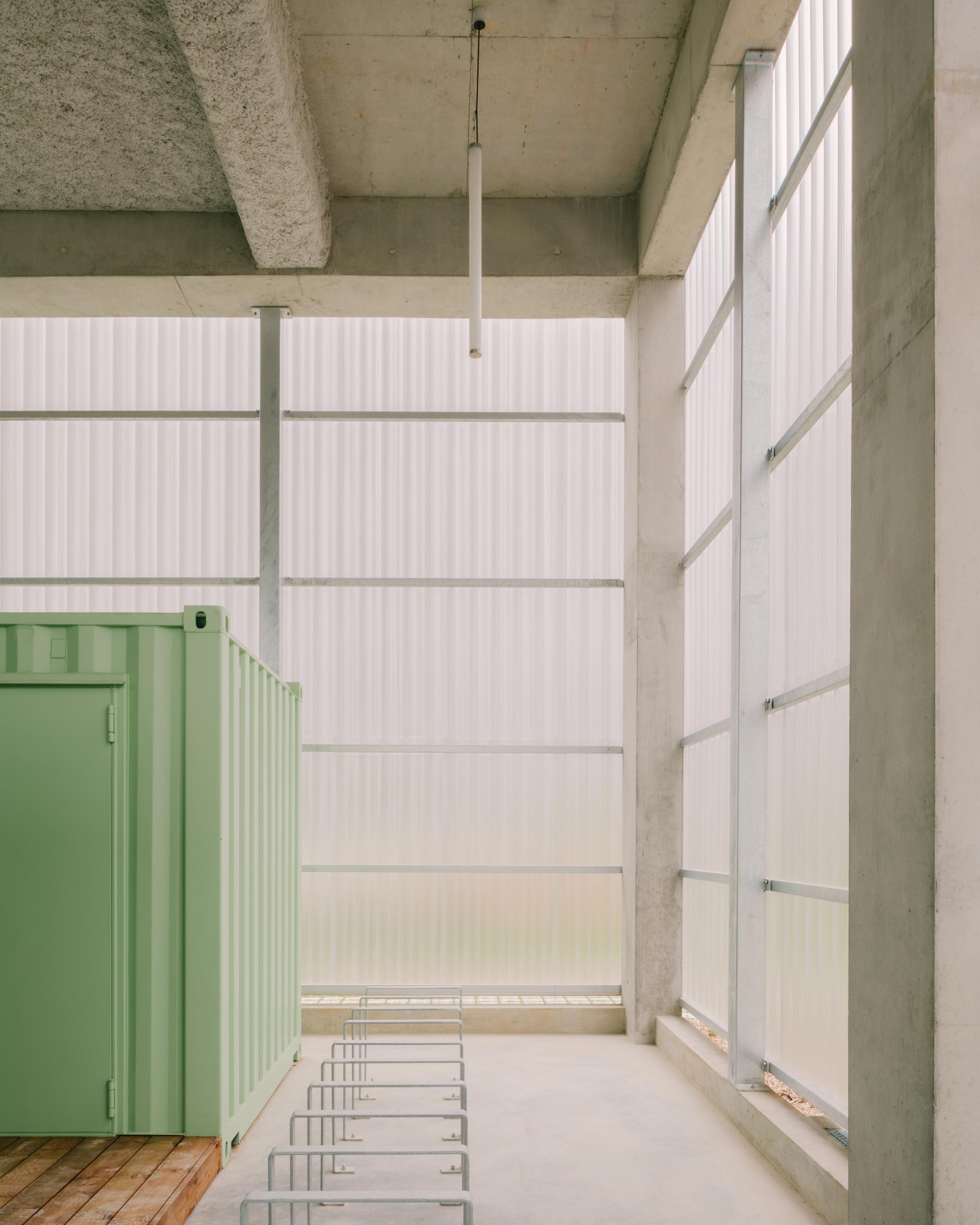 Vue intérieure des 116 logements à Vénissieux : structure béton, façades translucides, container vert technique et râteliers vélos sous lumière diffuse.