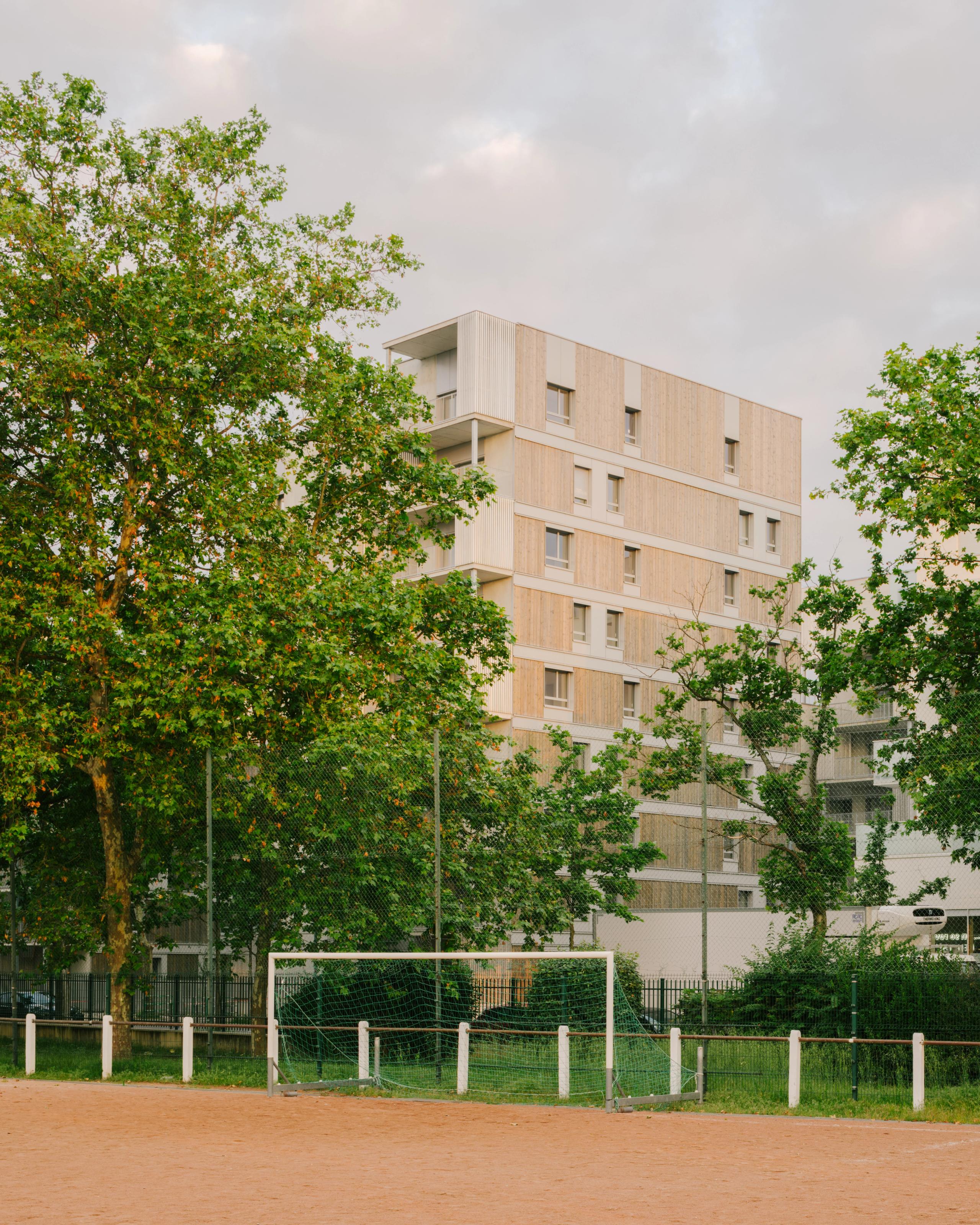 Vue du projet de 116 logements à Vénissieux : volume fragmenté en ossature bois et béton, façades rythmées, balcons en retrait, en lisière d’un terrain de sport arboré.