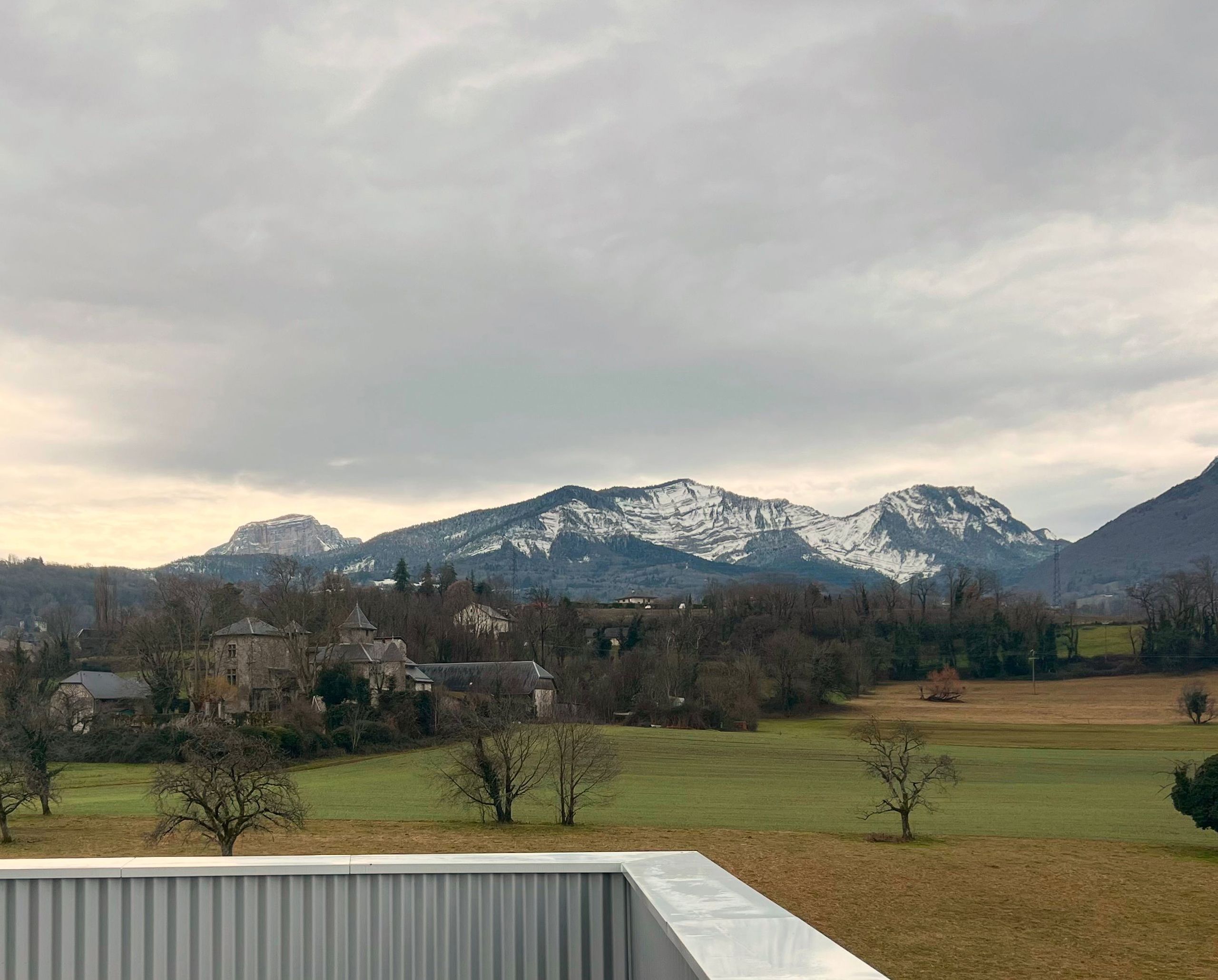 Vue depuis le projet de 142 logements à Cognin : acrotère métallique en premier plan, paysage rural et massif montagneux enneigé en arrière-plan.