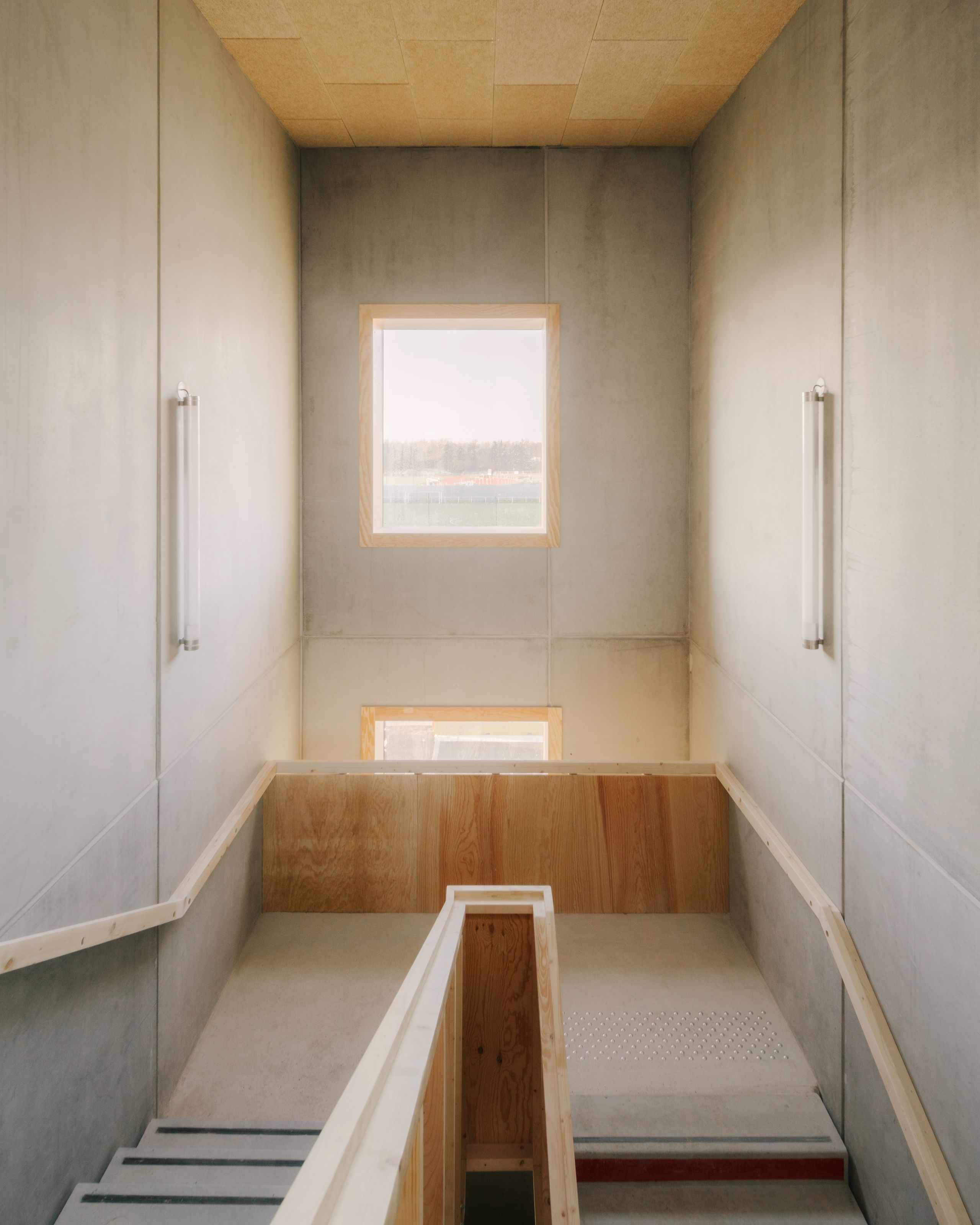 Escalier intérieur des groupes scolaires Jacqueline Auriol et Jean Jaurès au Bourget, cage en béton brut et bois clair, éclairée par une baie carrée.