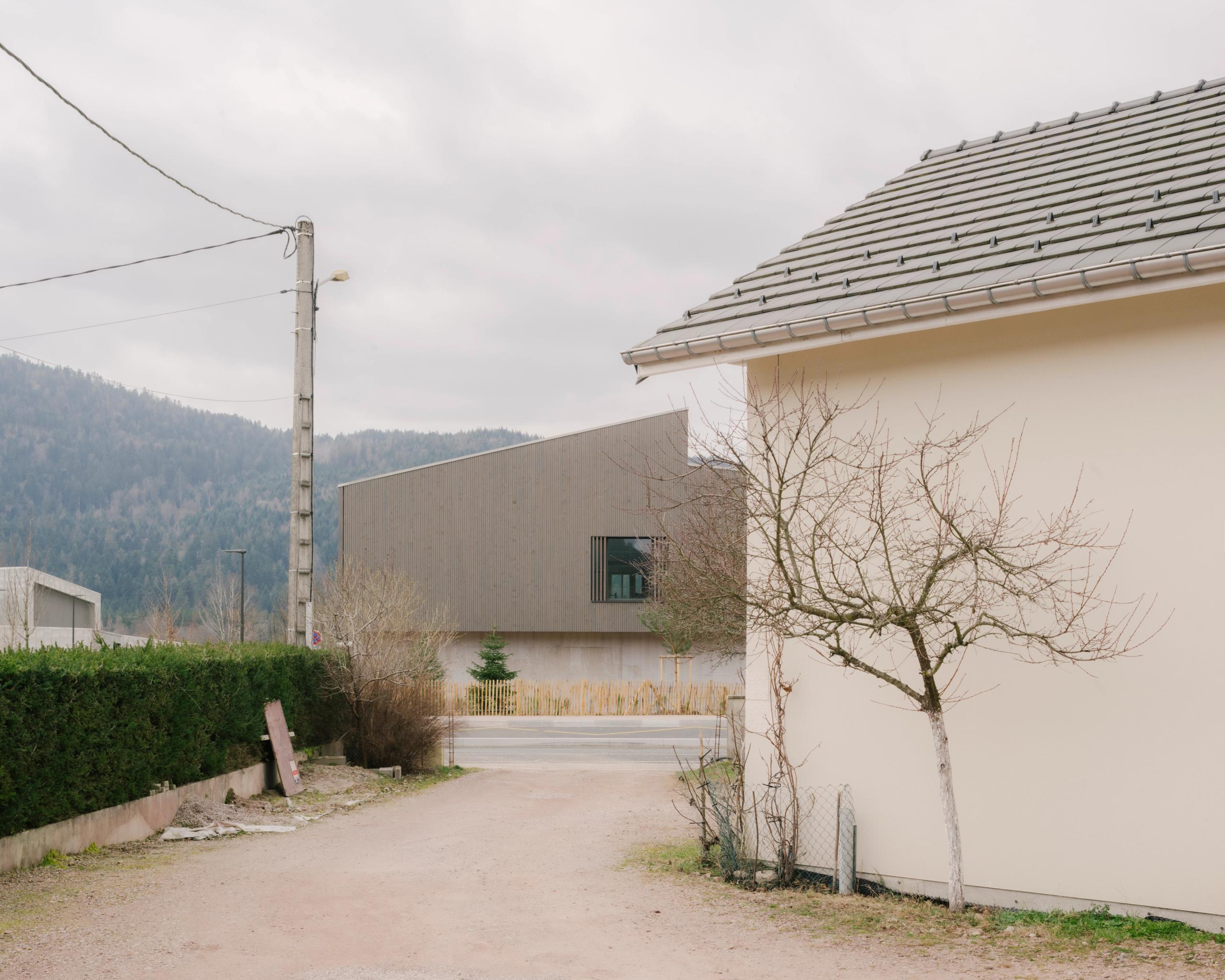 Vue lointaine du Collège du Ban à Vagney : volume en bois à toiture plate, façade rythmée par un bandeau vitré, inséré dans un paysage pavillonnaire vosgien.