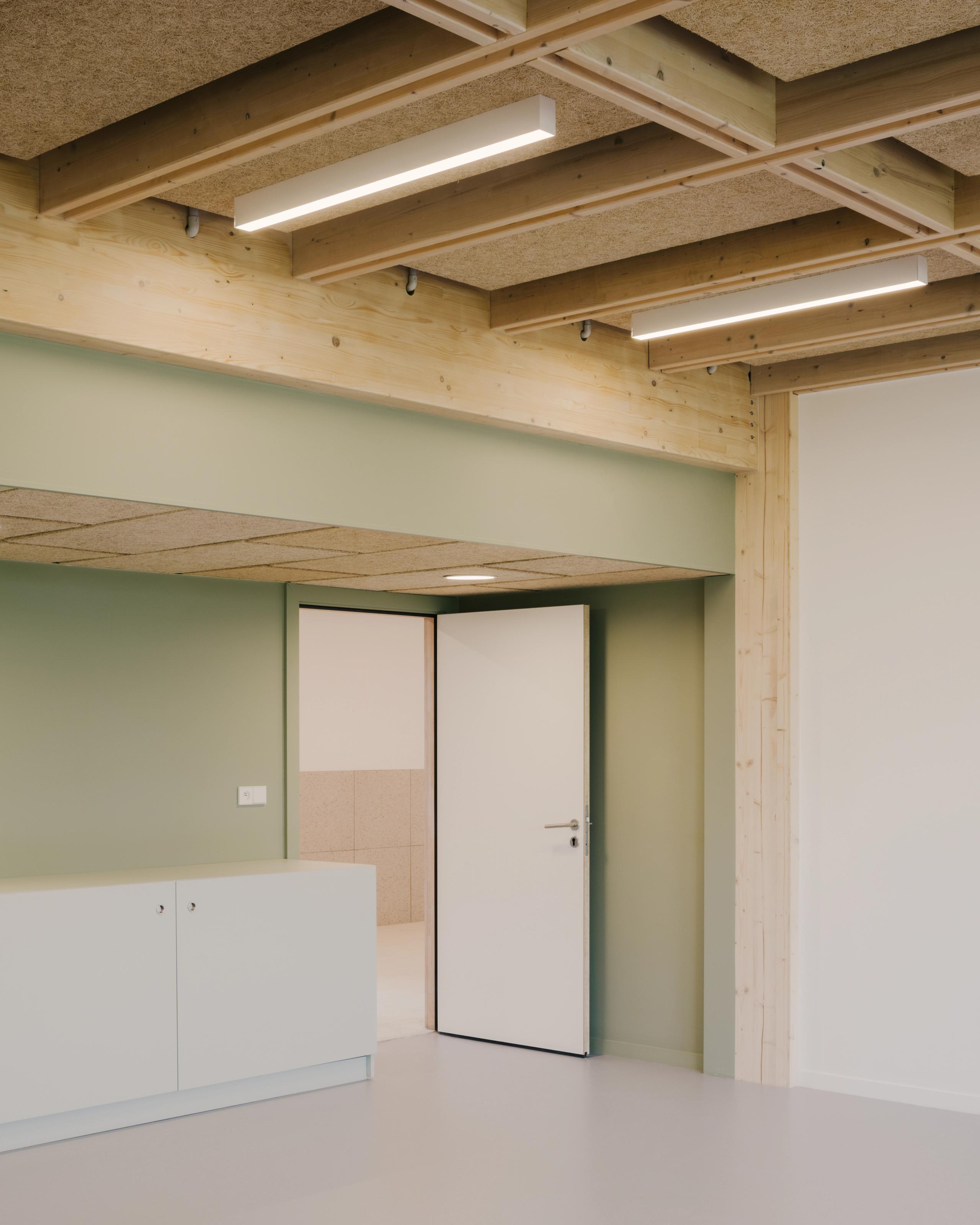 Intérieur du collège Maryse Bastié à Dole : extension en bois apparent, plafond caisson, parois vert pâle et porte blanche cadrant un espace clair.
