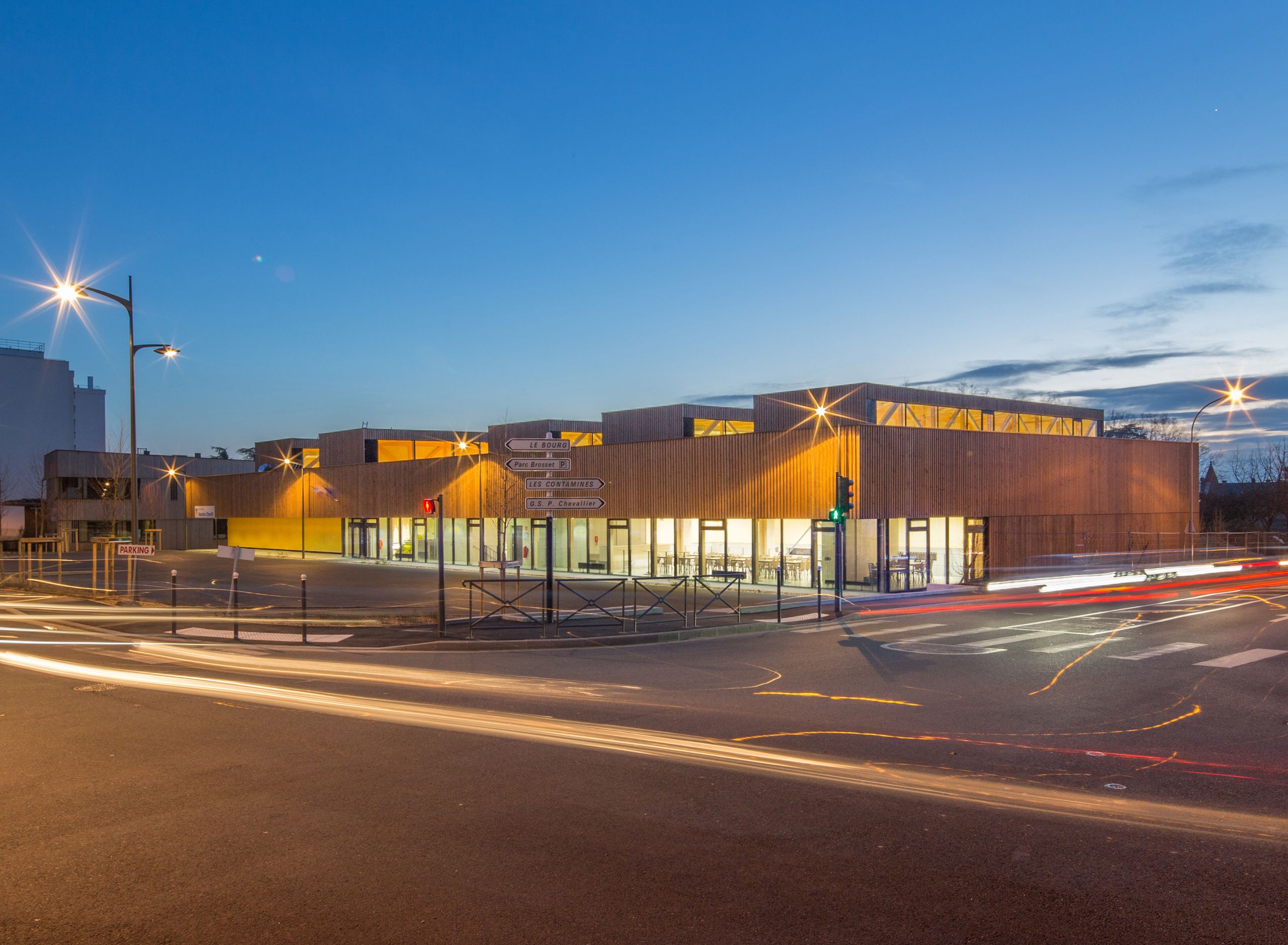Photographié au crépuscule, le gymnase Hacine Cherifi se présente comme un volume bas en bois à façade rythmée de lames verticales et de larges baies vitrées, ponctué de lanterneaux en toiture qui laissent filtrer une lumière chaude sur un carrefour urbain animé par les traînées lumineuses des voitures.