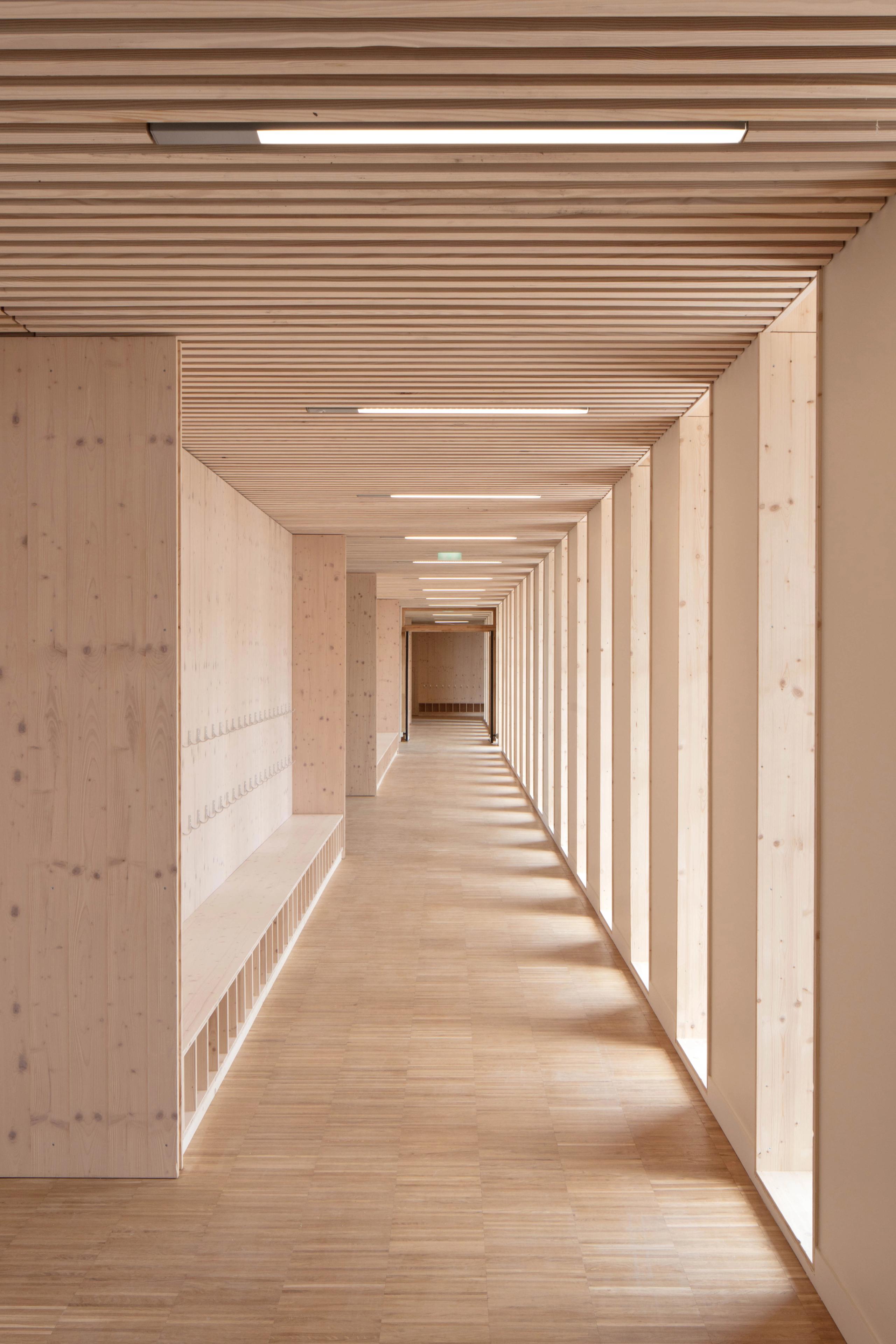 Couloir en bois du groupe scolaire Denis Mukwege à Melun, éclairé par une trame régulière de baies verticales et un plafond à lames avec luminaires intégrés.