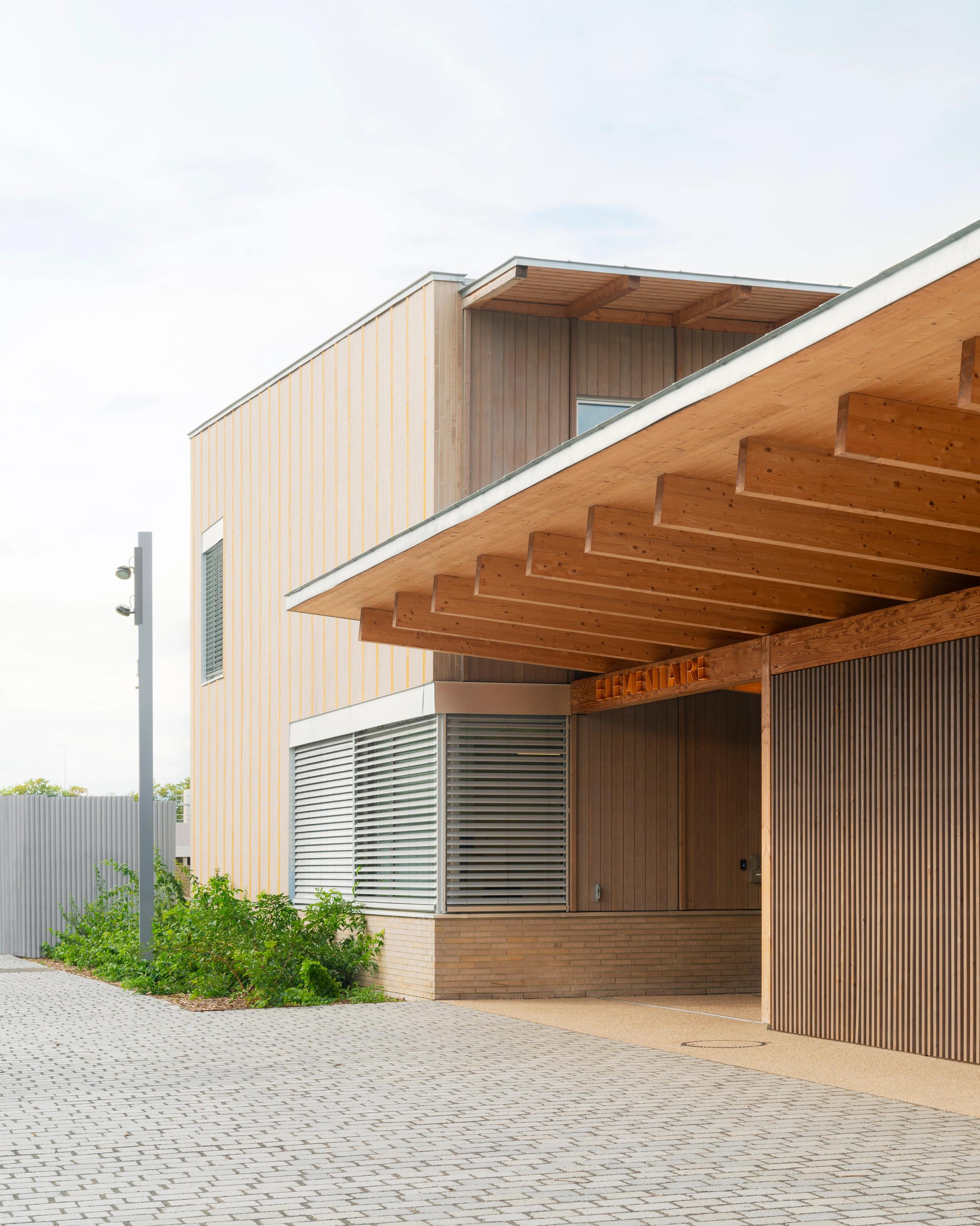 Façade des groupes scolaires Jacqueline Auriol et Jean Jaurès au Bourget : volumes en U en ossature bois, auvent saillant, brise-soleil métalliques et socle en brique.