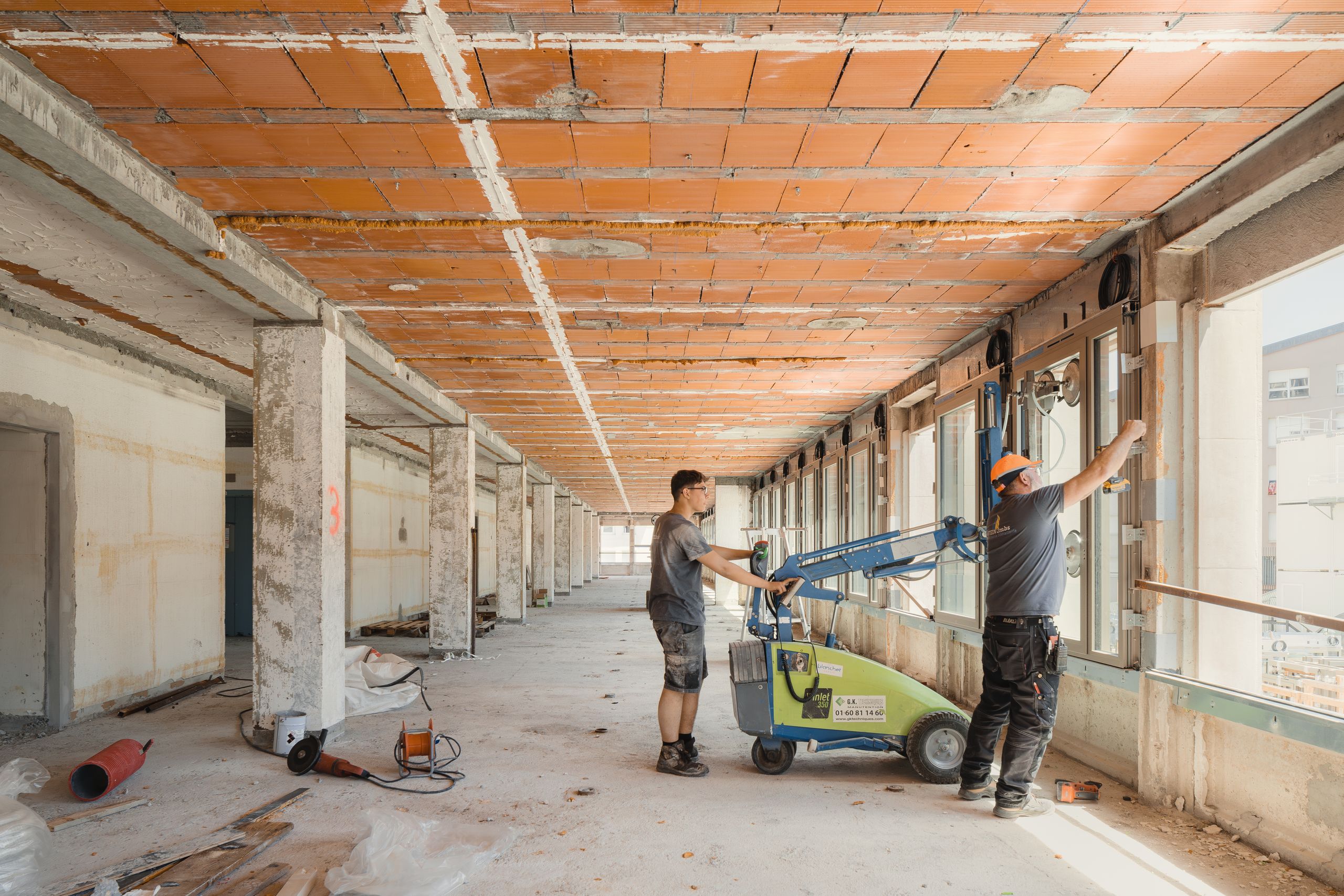 Chantier de réhabilitation de l’IGH Émile Loubet à Saint-Étienne, plateau nu en béton et briques avec façade vitrée en cours de dépose par deux ouvriers.