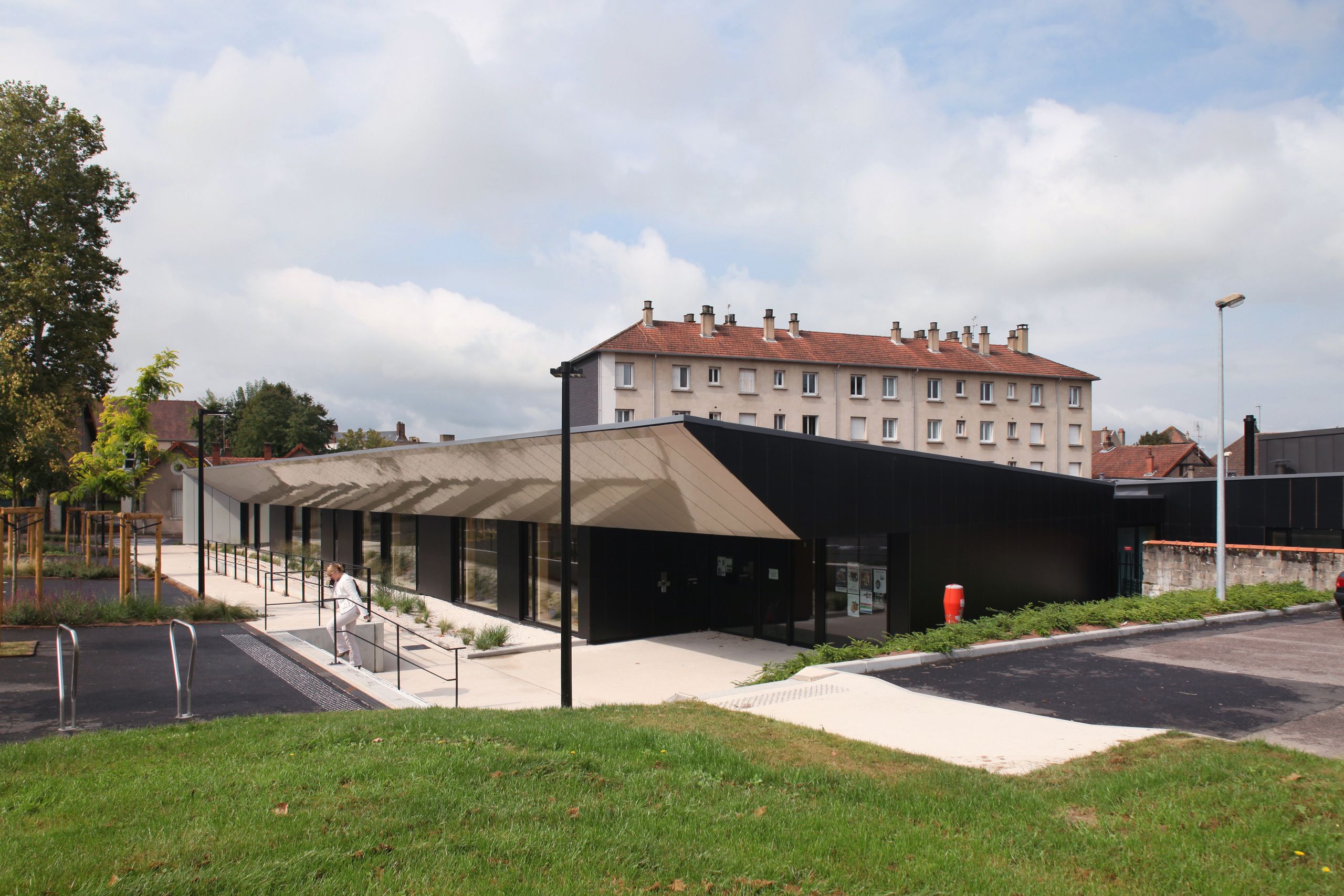 Maison de la petite enfance à Autun, volume en équerre noir et casquette métallique irisée, large galerie vitrée sur parvis minéral et jardin en pente.