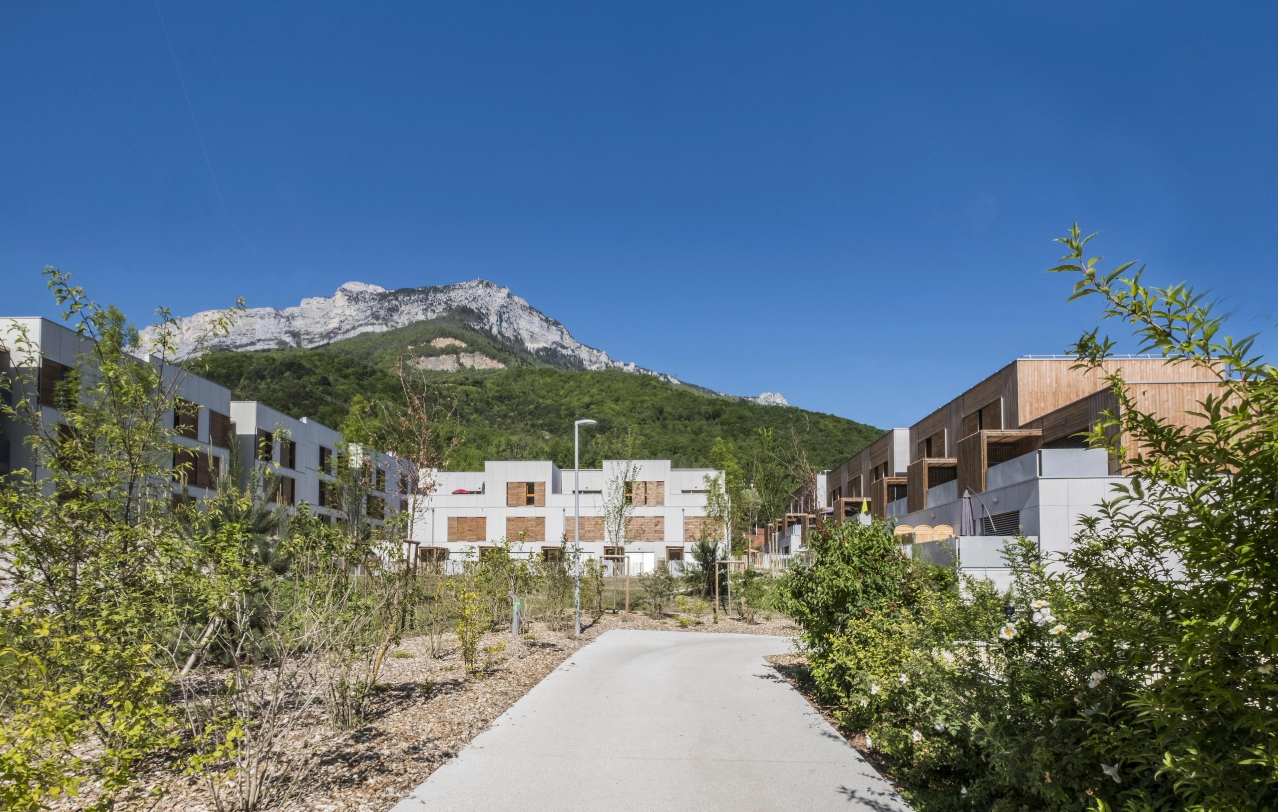 Vue des 65 logements à Seyssins par Tectoniques : volumes intermédiaires en béton et bois, chemin piéton central, jardin partagé, fond de massif du Vercors.