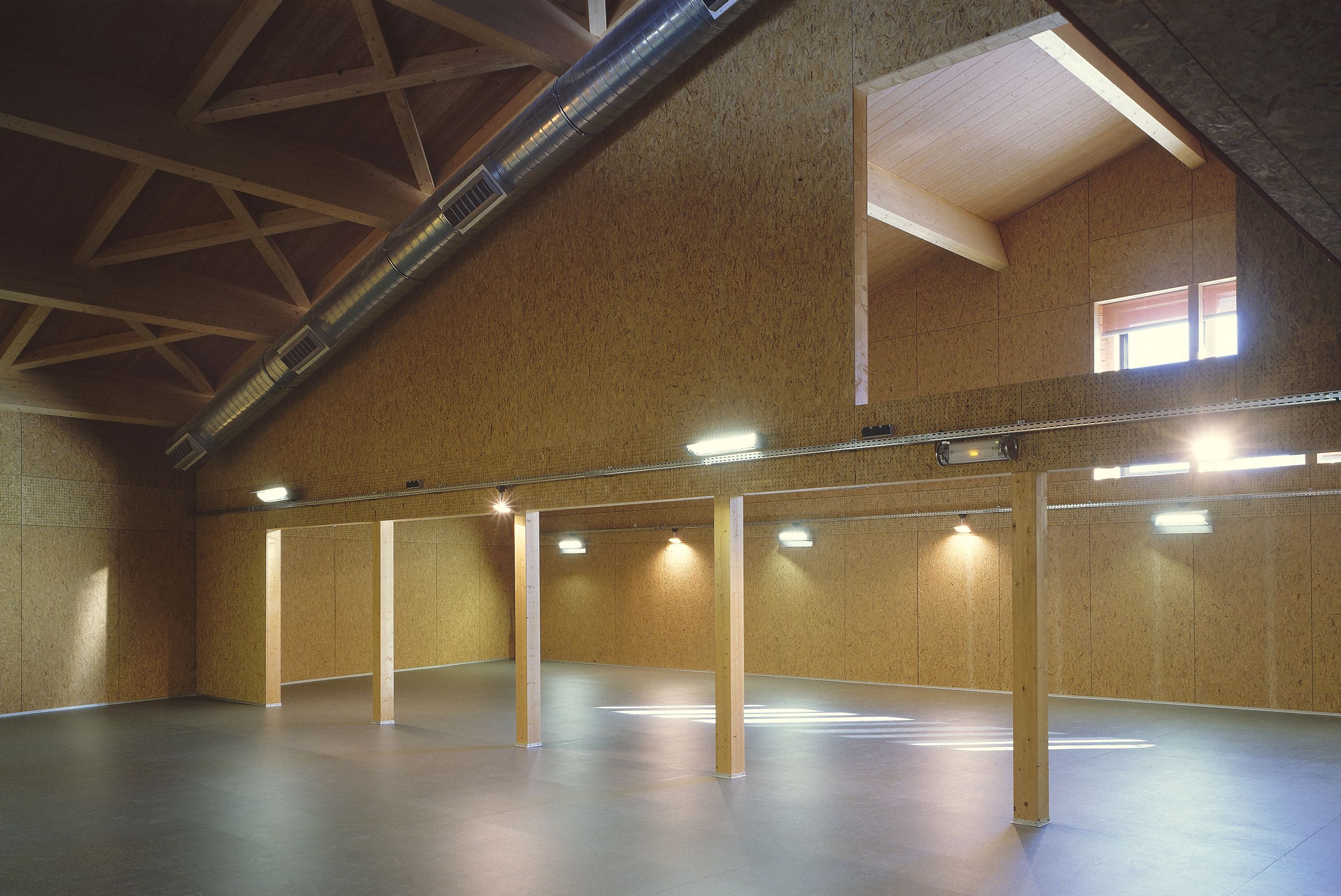 Intérieur de la Maison du Parc du Haut-Jura à Lajoux : grand volume en structure bois apparente, parois en panneaux agglomérés et gaines métalliques en sous-face de toiture.