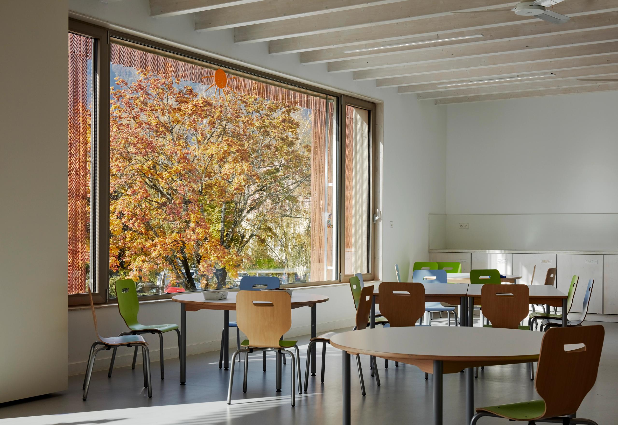 Interior view of Simone Veil Early Childhood Center in Albertville, with exposed beams, child‑scale tables and chairs, and a large glazed opening framing autumn trees.