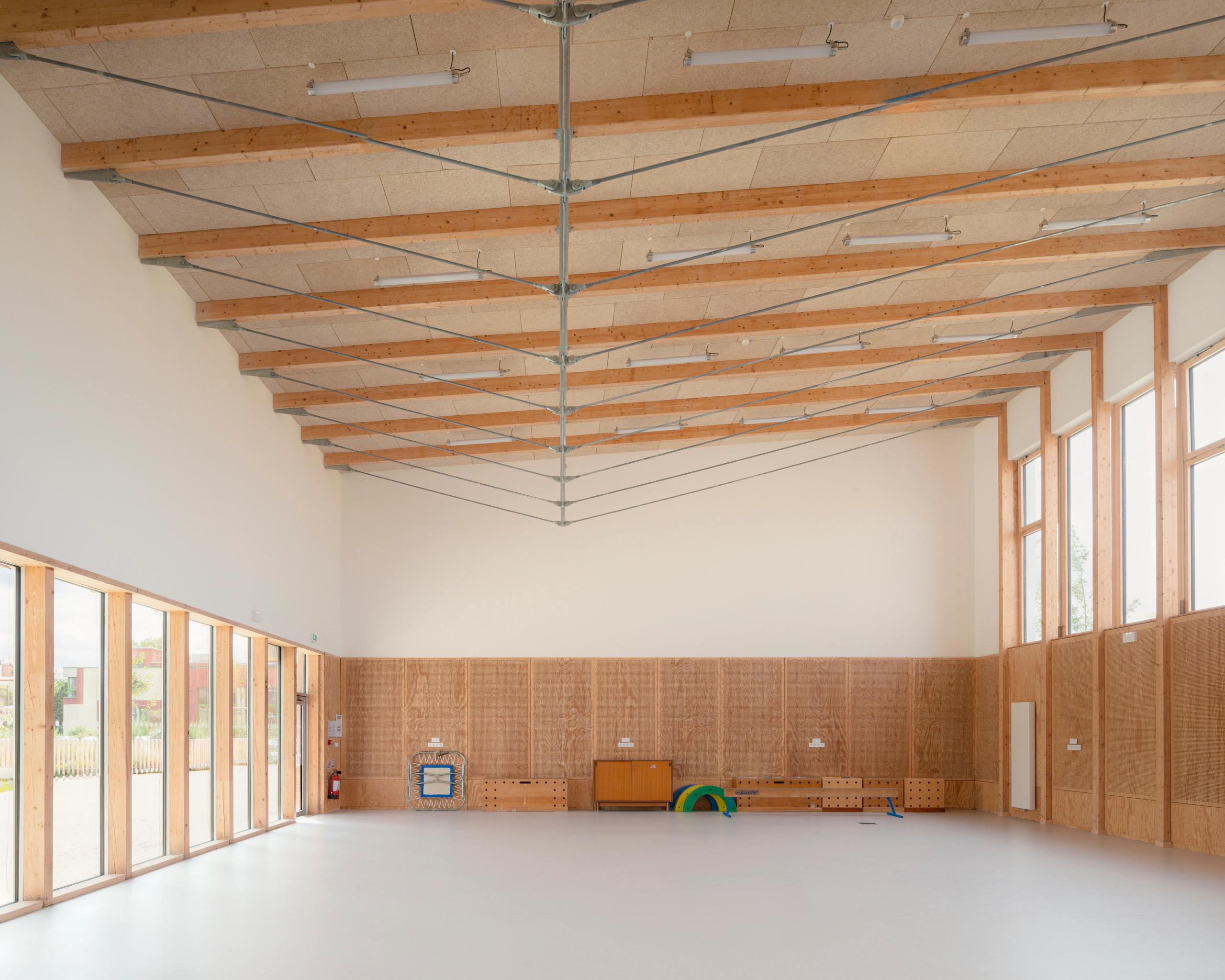 Grande salle des groupes scolaires Jacqueline Auriol et Jean Jaurès au Bourget, structure bois apparente, parois vitrées et panneaux de contreplaqué en soubassement.
