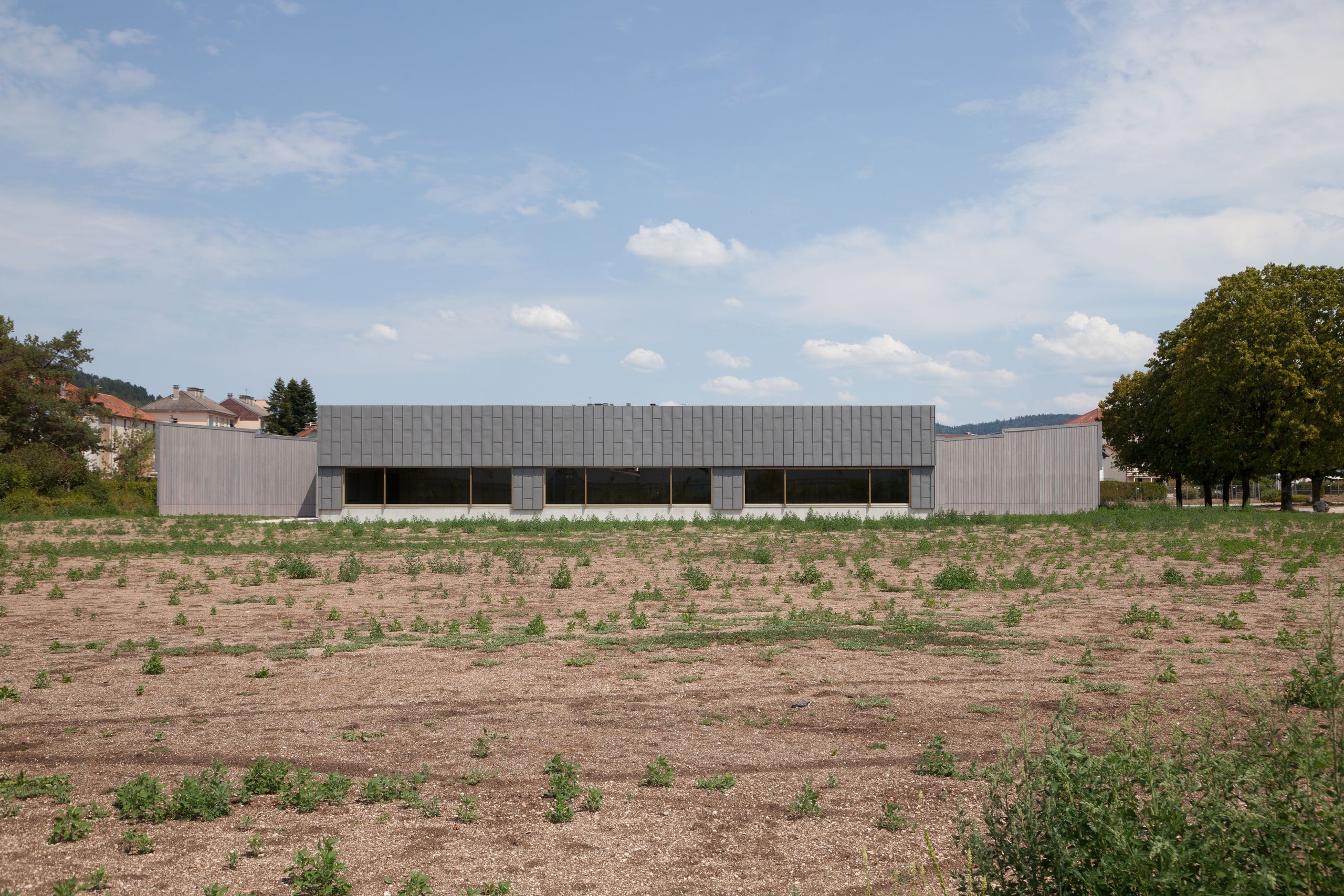 Vue lointaine de l’école élémentaire Hubert Reeves à Champagnole, volume bas en zinc et bois, façade horizontale vitrée ouvrant sur un terrain en friche.
