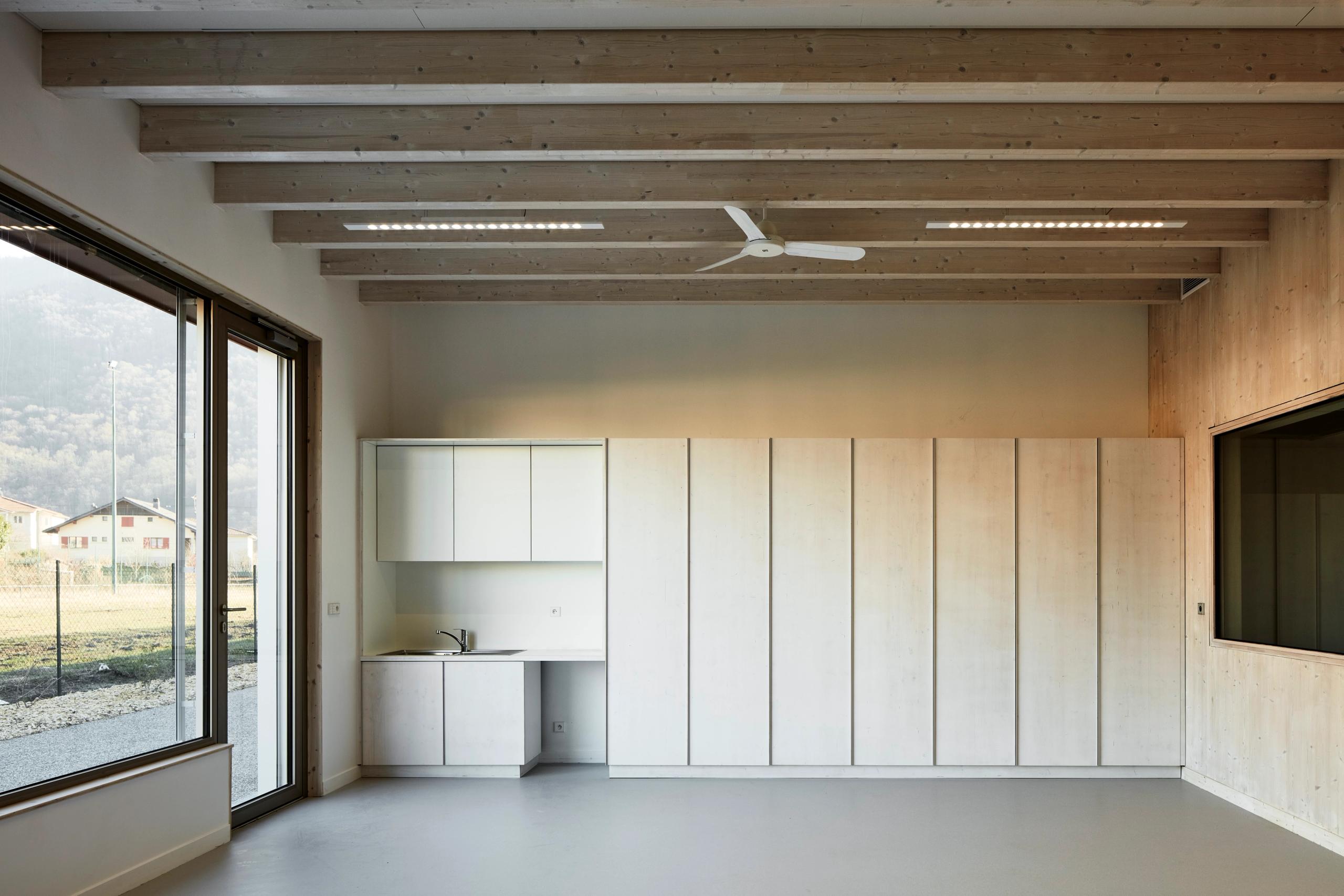 Intérieur de la Maison de l’Enfance Simone Veil à Albertville : salle lumineuse en bois apparent, grands rangements intégrés, kitchenette et baie vitrée sur le paysage alpin.