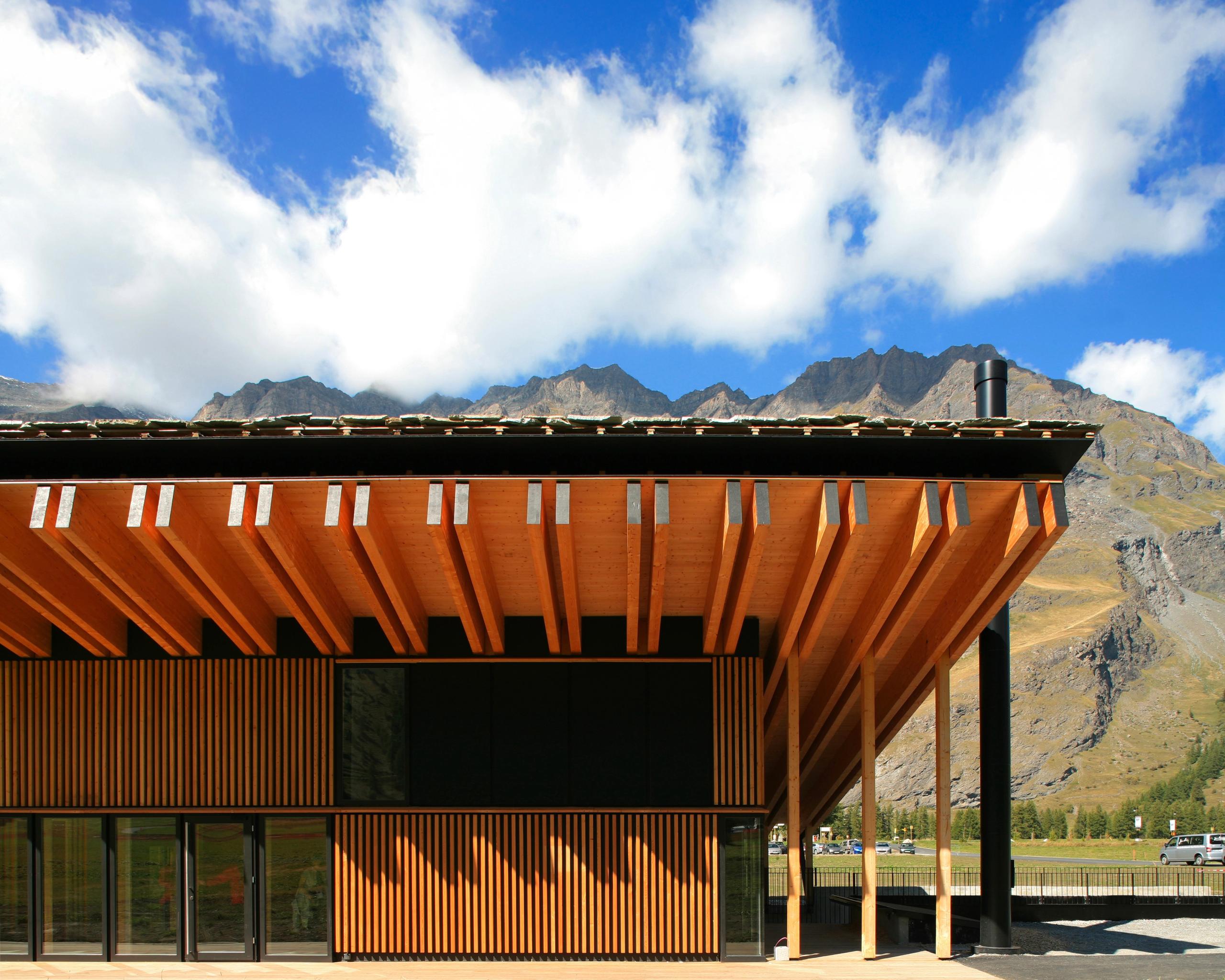 Espace nordique de Bessans : volume en bois à portiques saillants, bardage à claire-voie et toiture en lauze, implanté face au paysage montagneux ouvert.