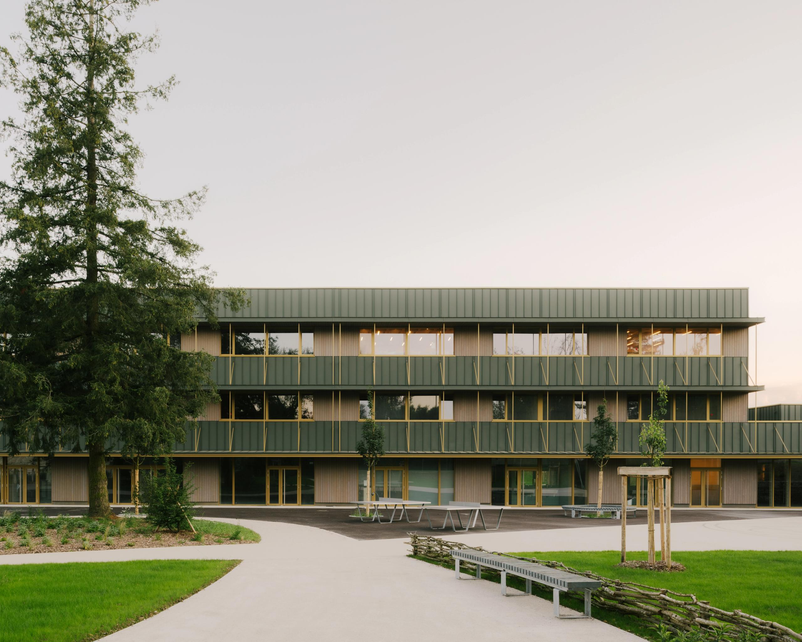 Façade restructurée du collège Maryse Bastié à Dole : volumes horizontaux bois-métal vert, trame rationnelle de baies vitrées, parvis minéral et jardins plantés.