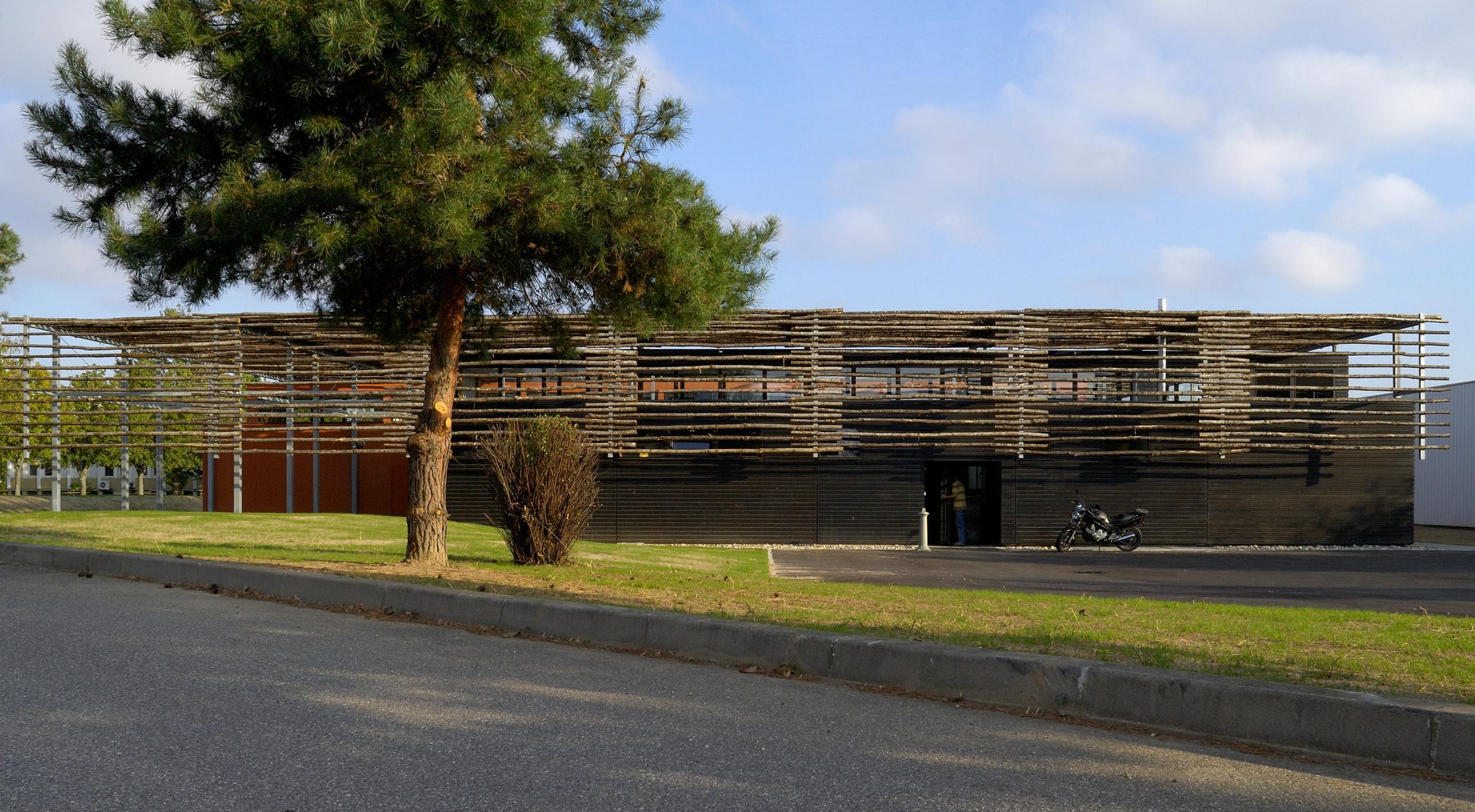 Façade du restaurant administratif du CETE à Bron, volumes bas en bois sombre sous ombrière de baliveaux de châtaignier, trame régulière et terrasse abritée.