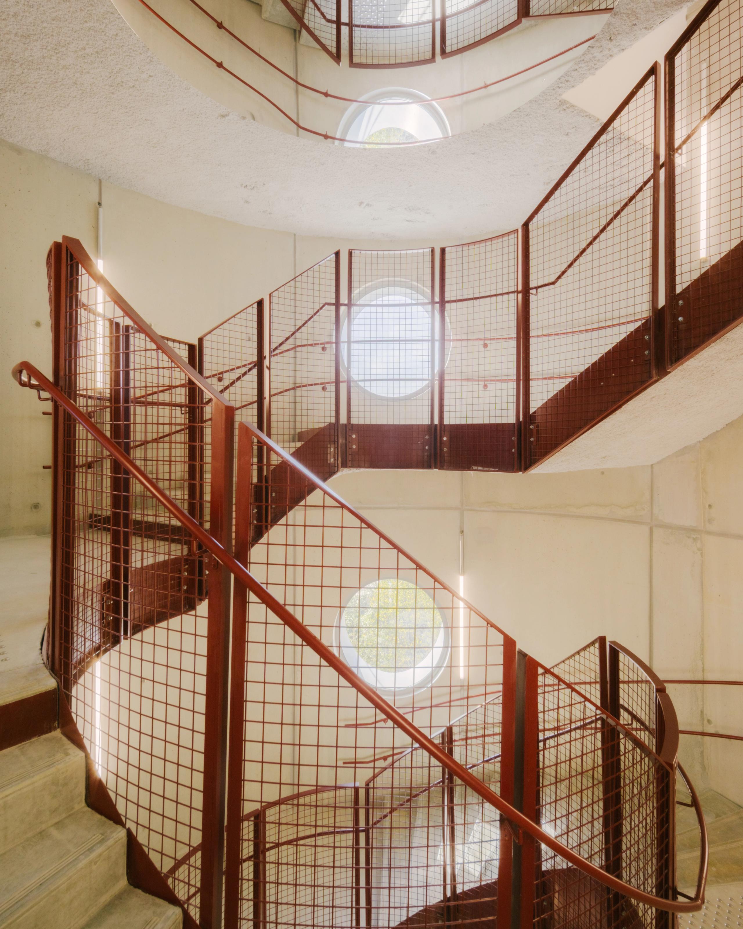 Escalier hélicoïdal du Groupe scolaire Dominique Frelaut à Colombes, garde-corps métalliques rouges et percements circulaires dans une cage en béton clair.