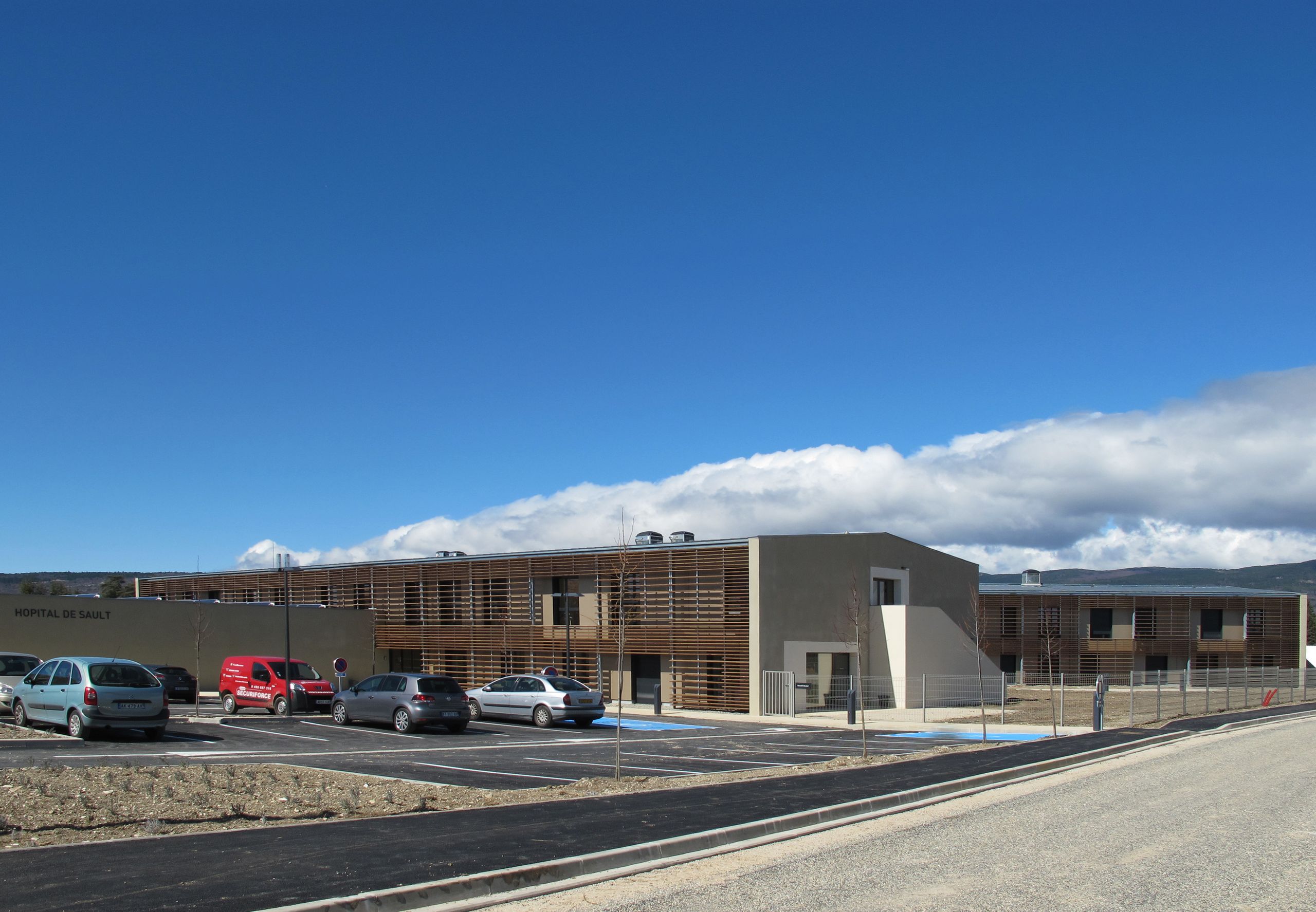 Vue de l’EHPAD de l’Hôpital de Sault : bâtiment bas en H, façades enduites et brise-soleil bois, larges baies vitrées ouvrant sur parkings et paysage.