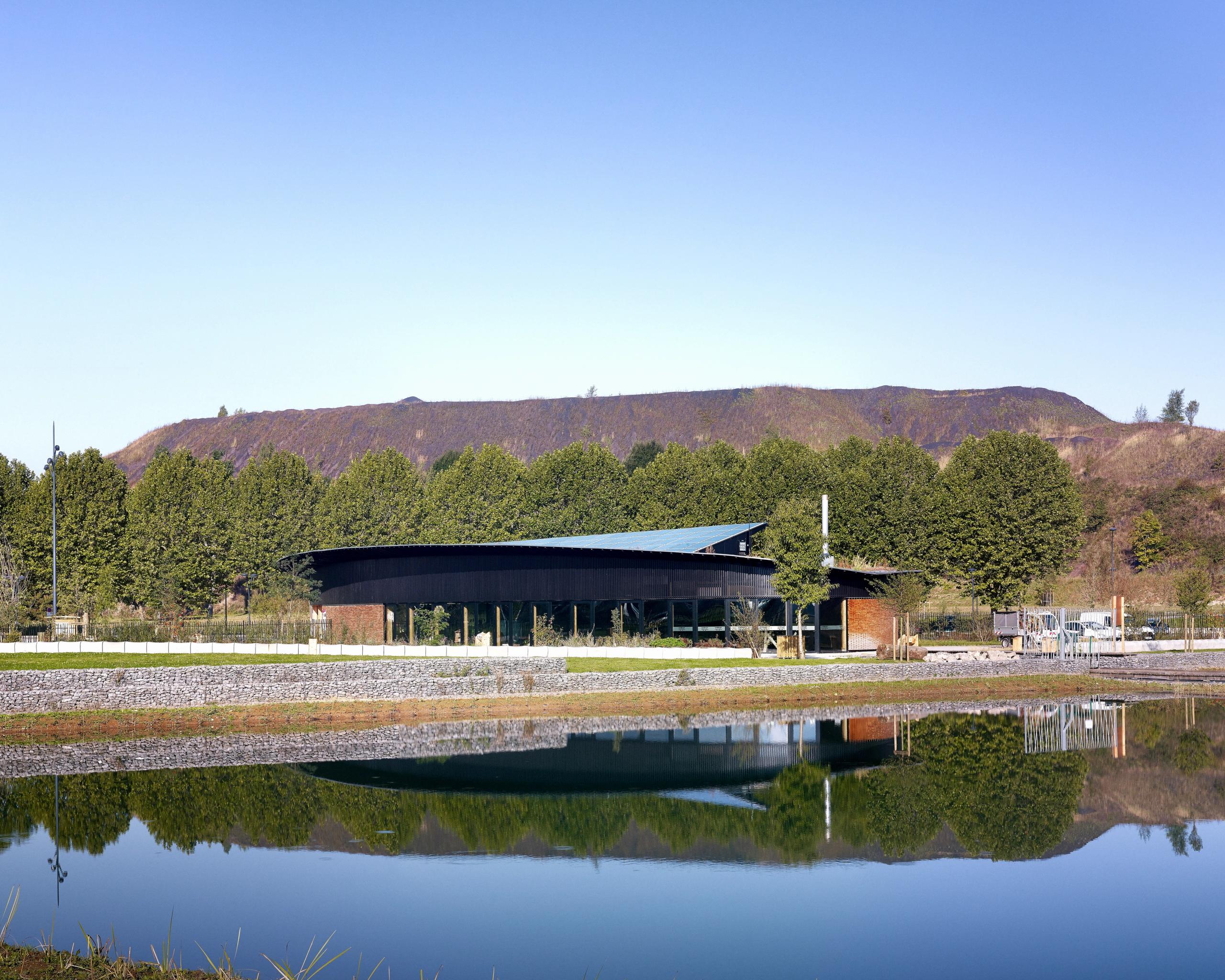 Maison de l’Environnement à Hénin-Beaumont, pavillon ovale en ossature bois sombre au bord d’un plan d’eau, intégré au paysage d’un ancien site minier.
