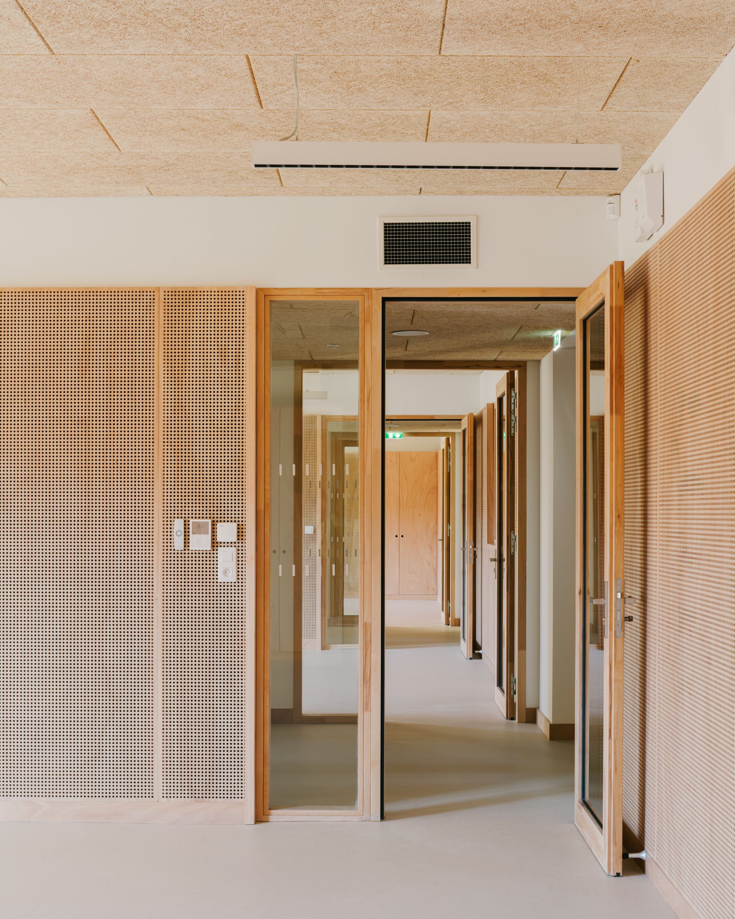 Couloir du pôle culture et jeunesse à Vaison-la-Romaine, rythmé par des portes vitrées en bois, parois acoustiques perforées et plafond en panneaux fibreux.