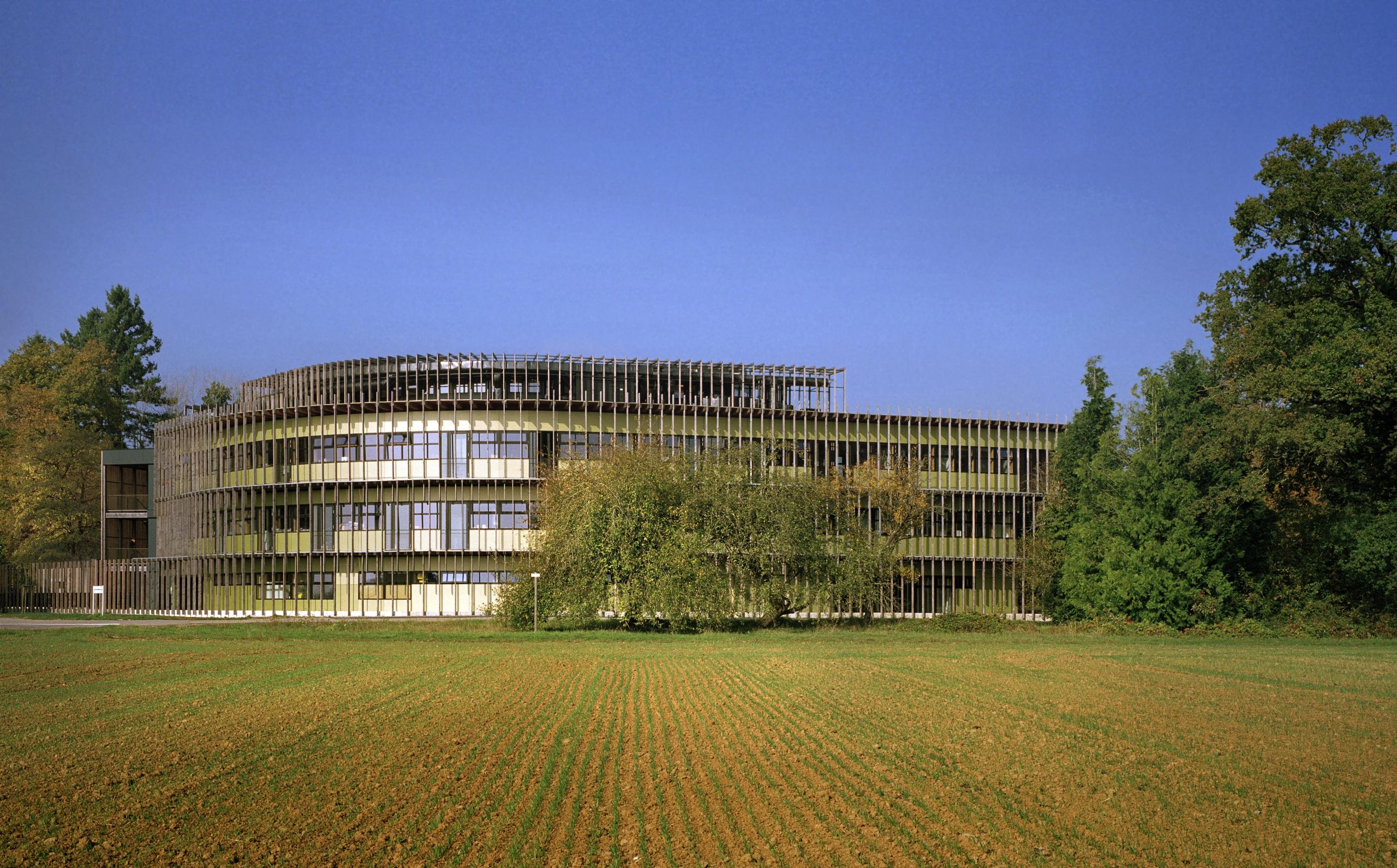 Façade sud courbe du laboratoire INRAE à Champenoux, enveloppe bois à lames verticales, grands vitrages continus, bâtiment inséré entre champ cultivé et lisière forestière.