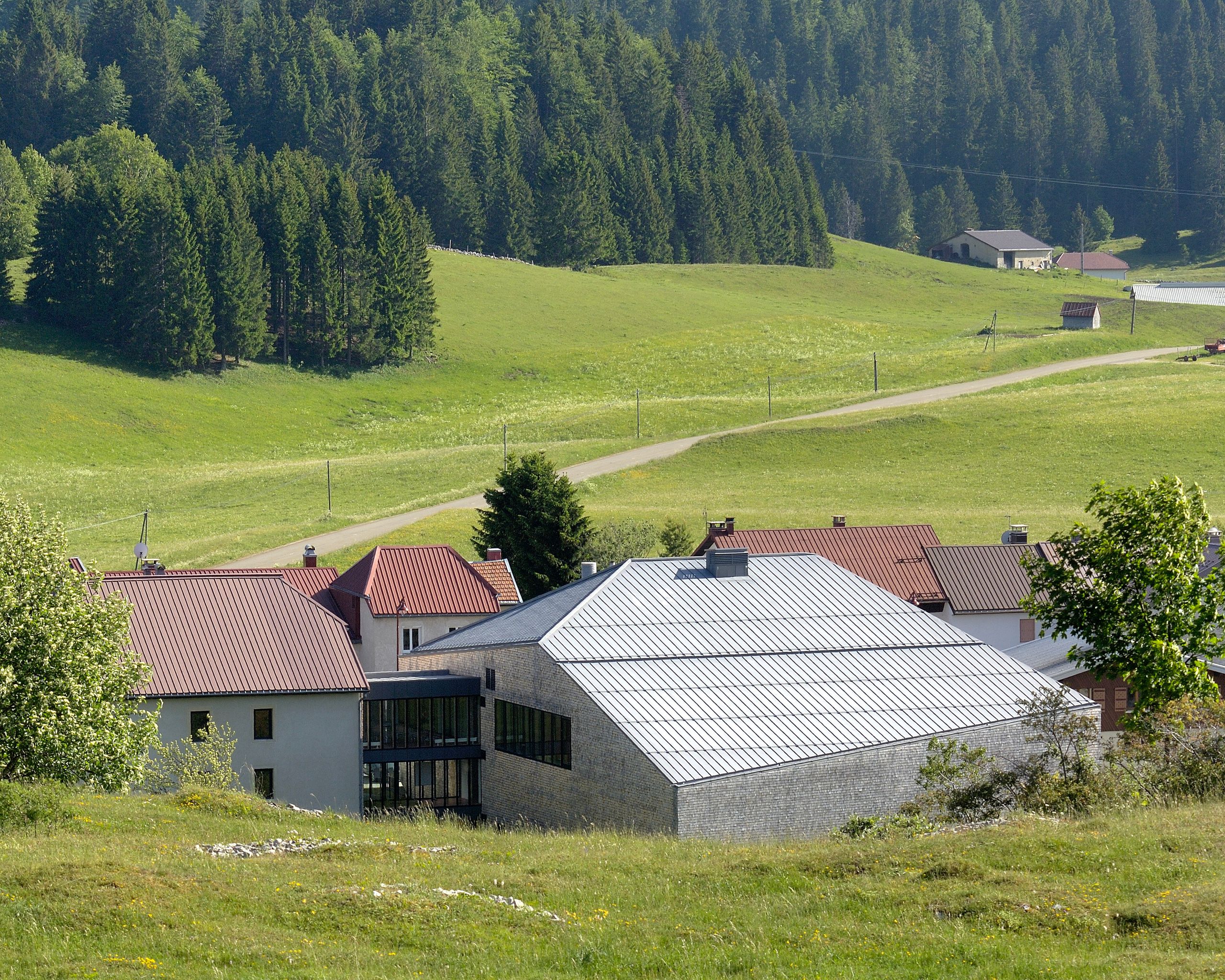 Extension de la Maison du Parc du Haut-Jura à Lajoux, volume compact en tavaillons d’épicéa et toiture métallique, inséré dans le village face aux prairies et forêts.