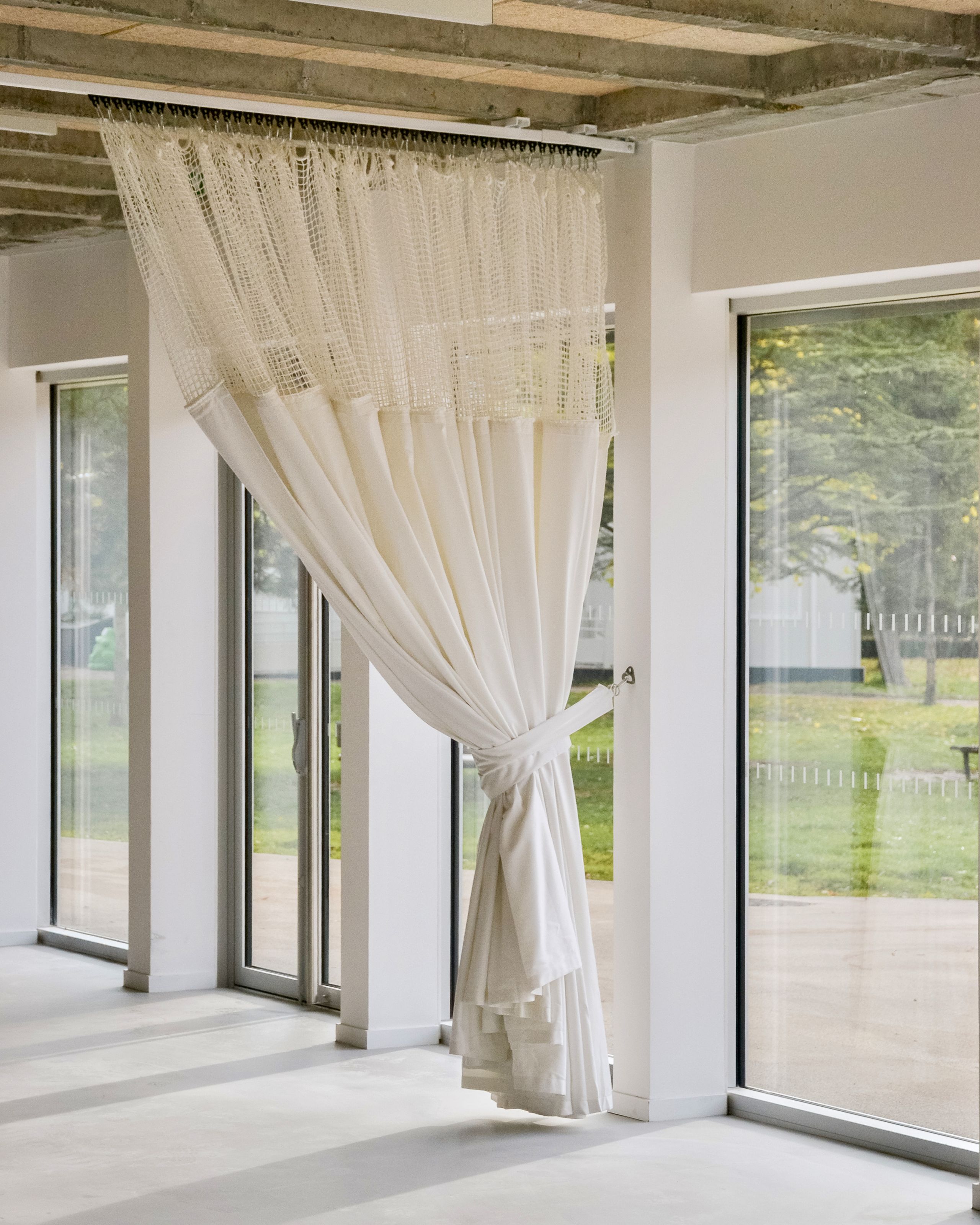 Intérieur du groupe scolaire du Bouchet à Bonneville : façade vitrée toute hauteur, rideau textile filtrant, structure béton apparente et sol clair continu.