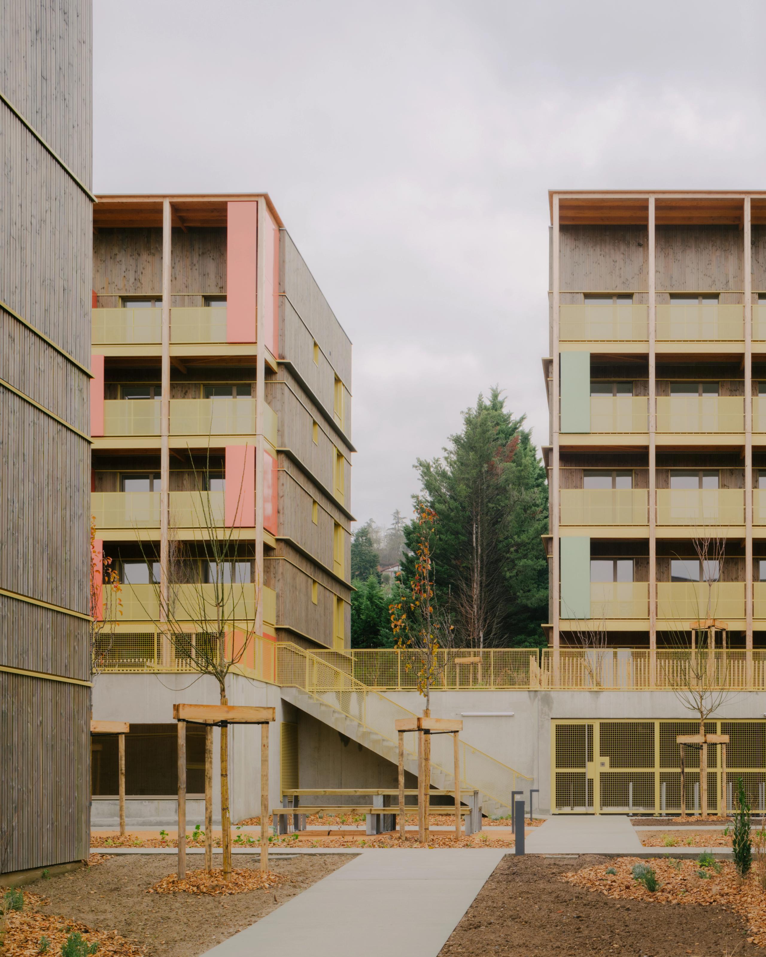 Vue des 84 logements Tectoniques à Trévoux : volumes en gradins bois-béton, coursives et balcons colorés, exo-structures bois cadrant une cour plantée.