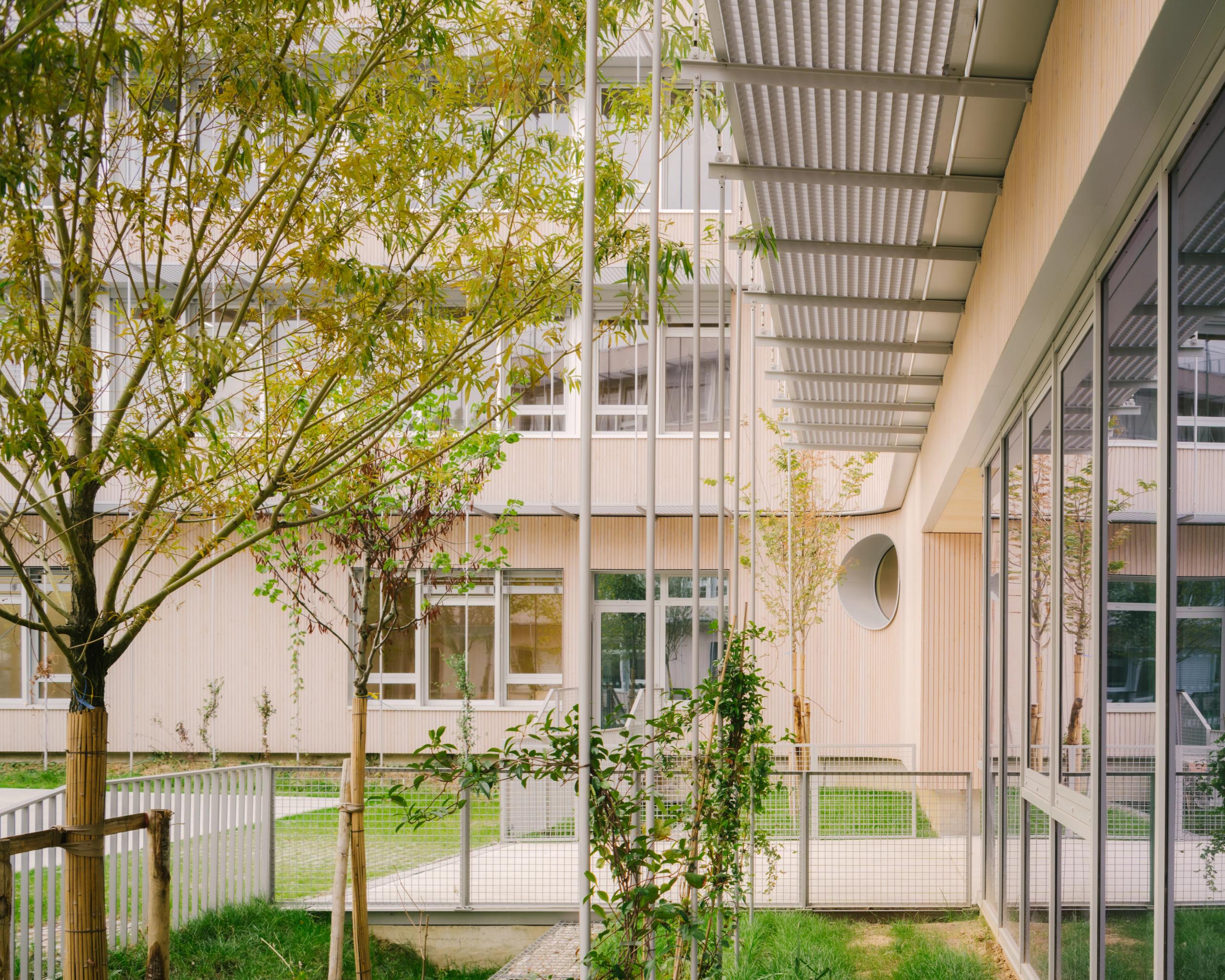 Patio planté du groupe scolaire Dominique Frelaut à Colombes, façades bois et menuiseries vitrées sous casquette métallique filtrant la lumière.