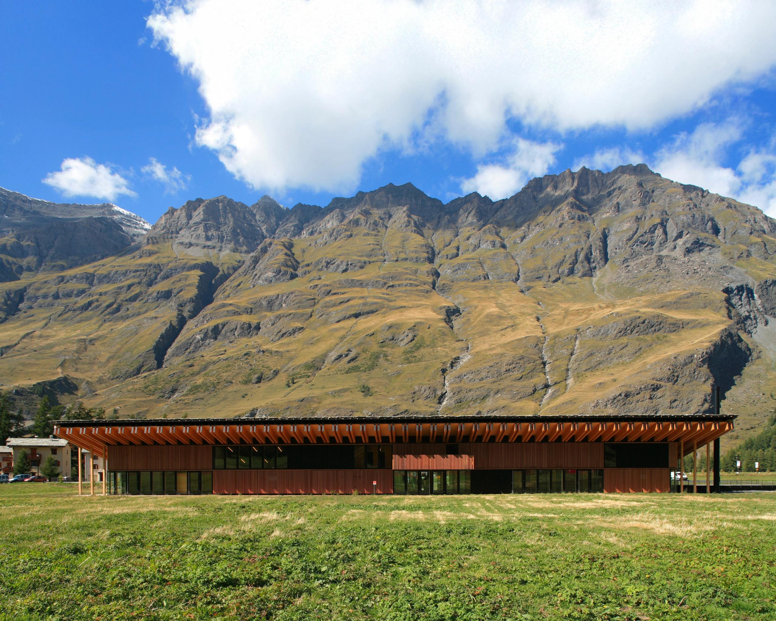 Espace nordique de Bessans : volume bas en bois et métal, toiture à large débord face aux pistes, implanté en lisière de prairie au pied du massif alpin.