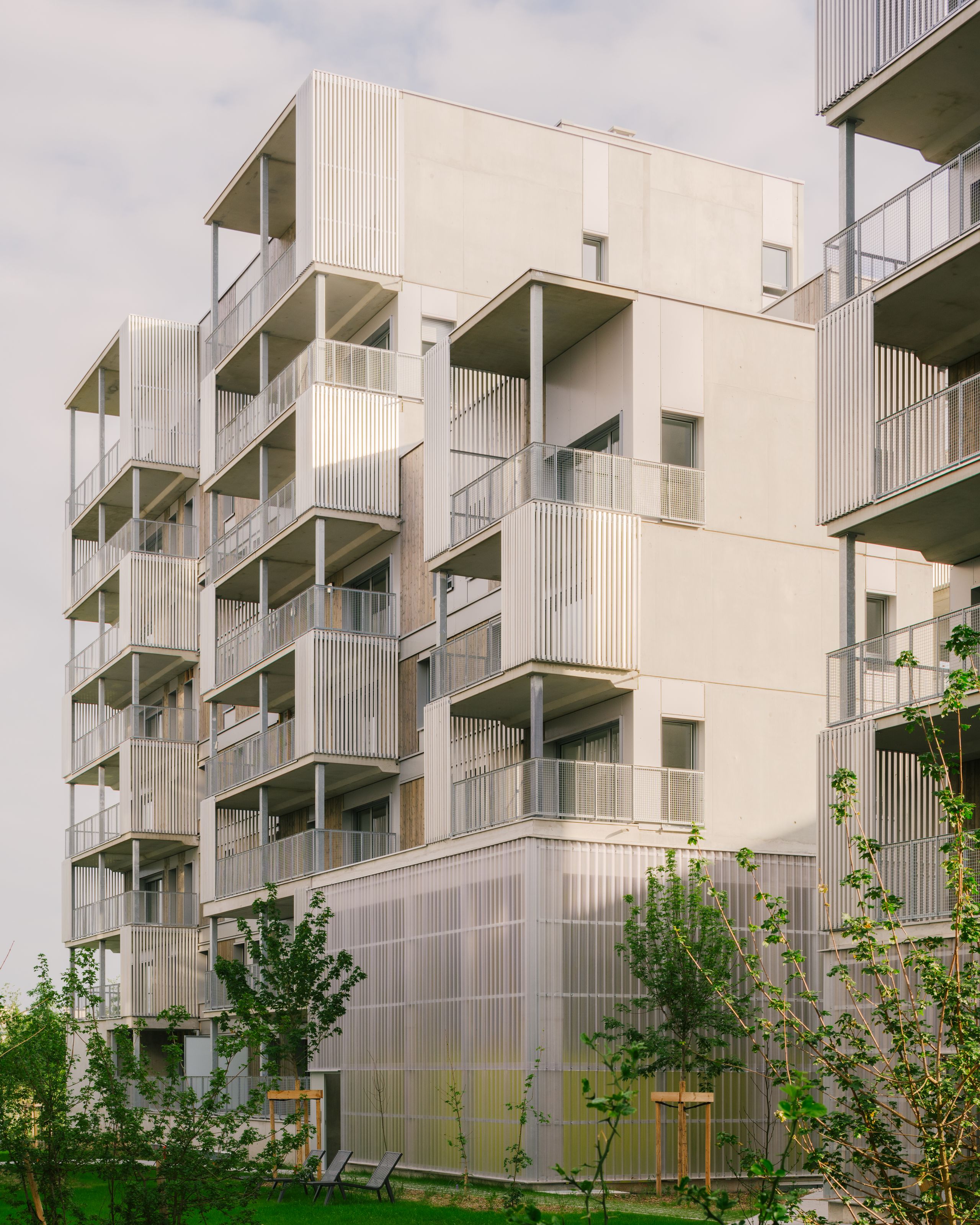 Vue des 116 logements à Vénissieux : volumes fragmentés, façades béton et ossature bois, balcons en exosquelette métallique sur jardin planté.