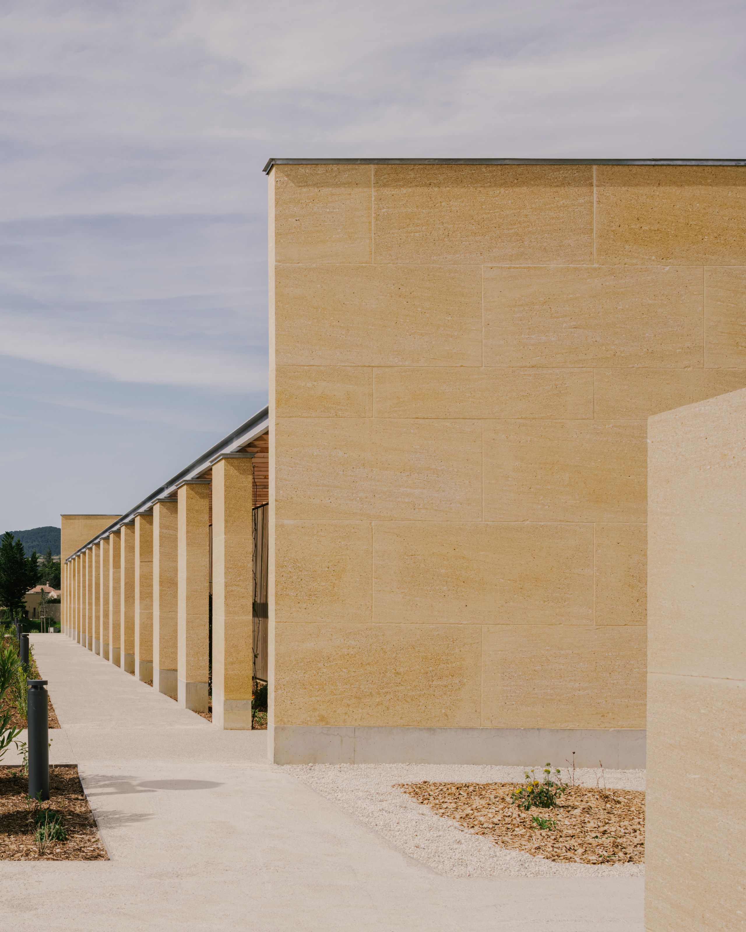 Youth Cultural Centre in Vaison-la-Romaine, showing a long stone enclosure with rhythmic piers, sheltered walkway and mineral forecourt under a clear sky.
