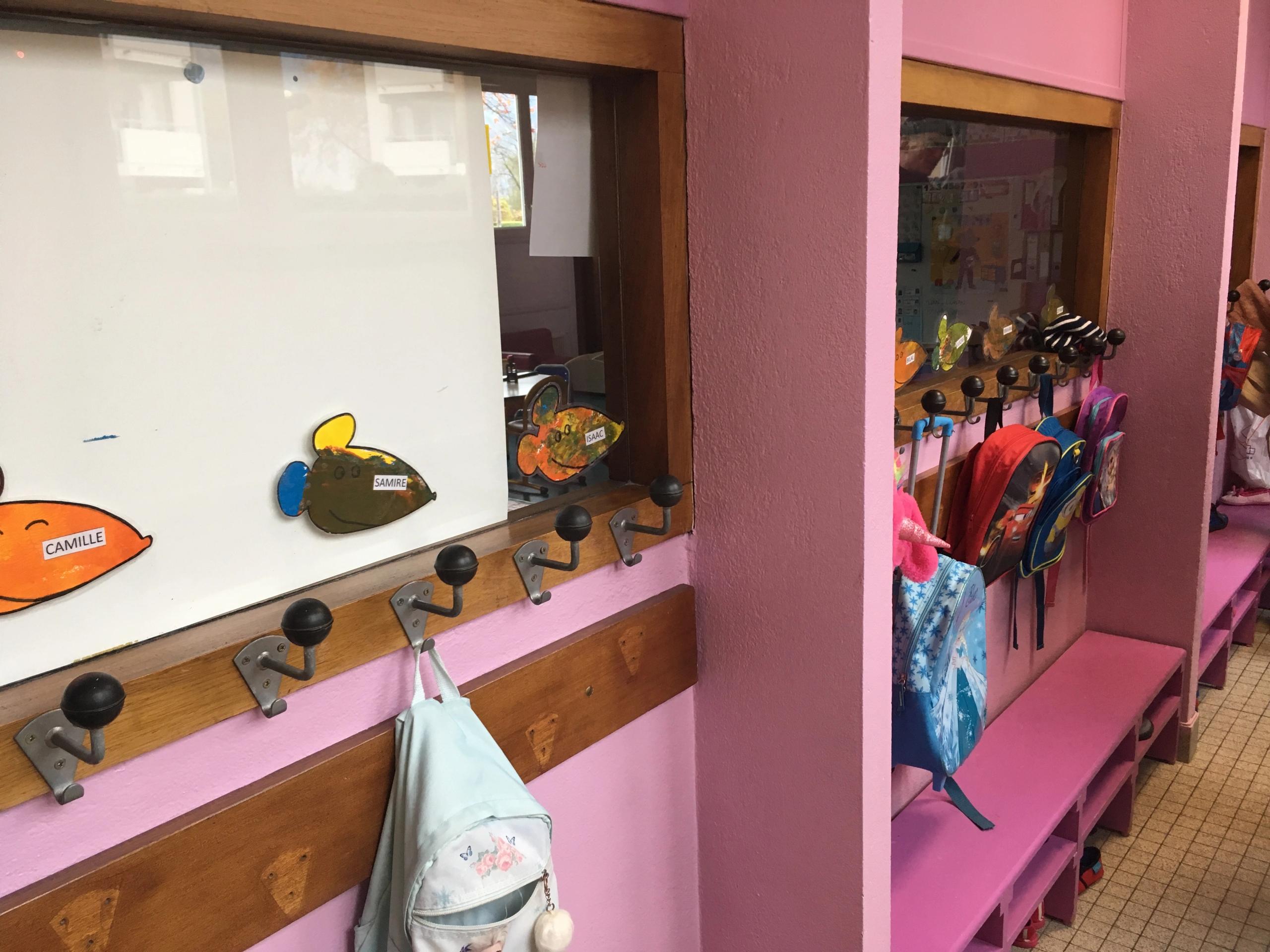 Vestiaire existant du groupe scolaire du Bouchet à Bonneville, couloir rose avec patères métalliques, bancs intégrés et rangements bas pour cartables d’élèves.