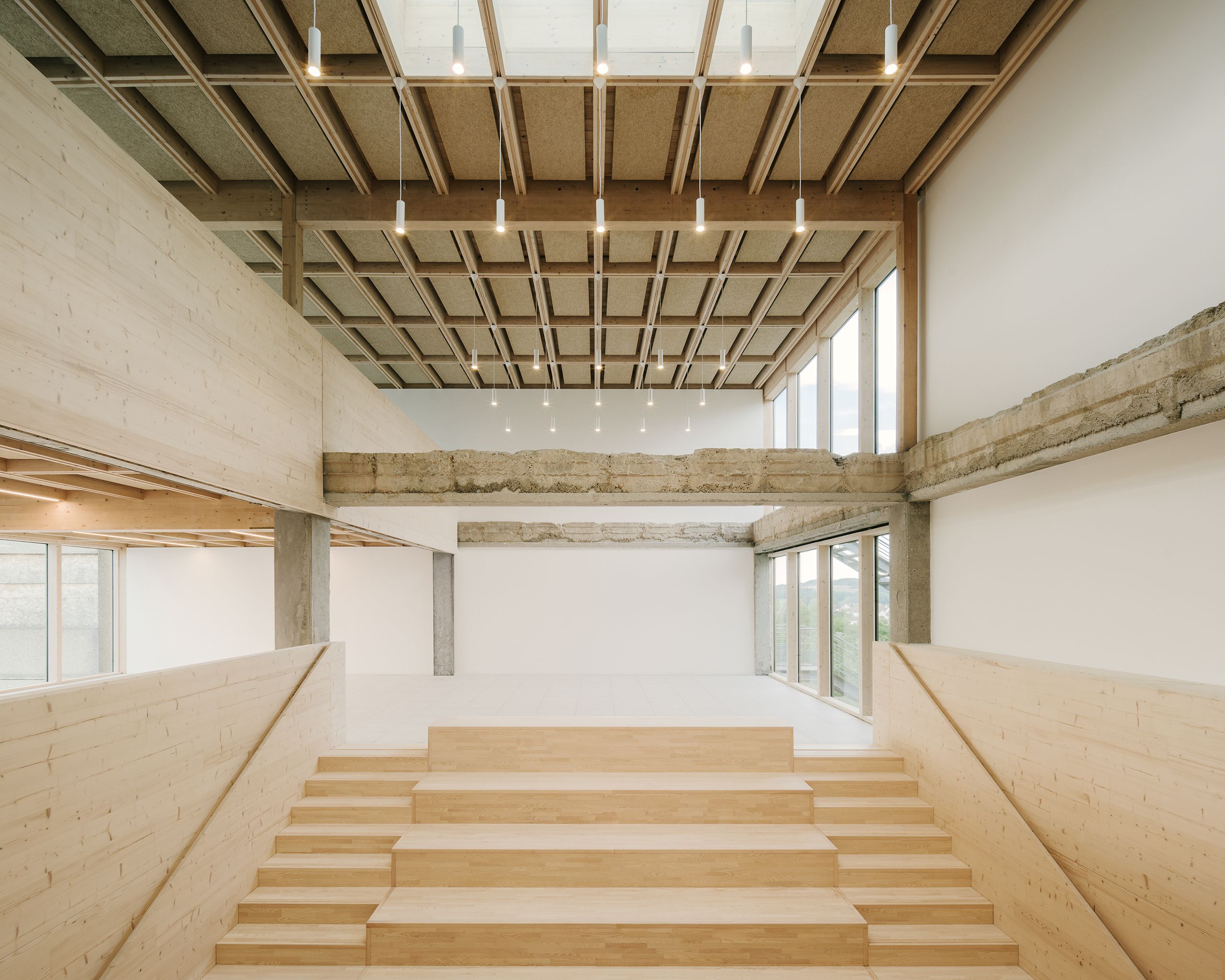 Espace intérieur du collège Maryse Bastié à Dole, gradins en bois clair, structure béton apparente et plafond caisson bois, largement ouvert sur l’extérieur.