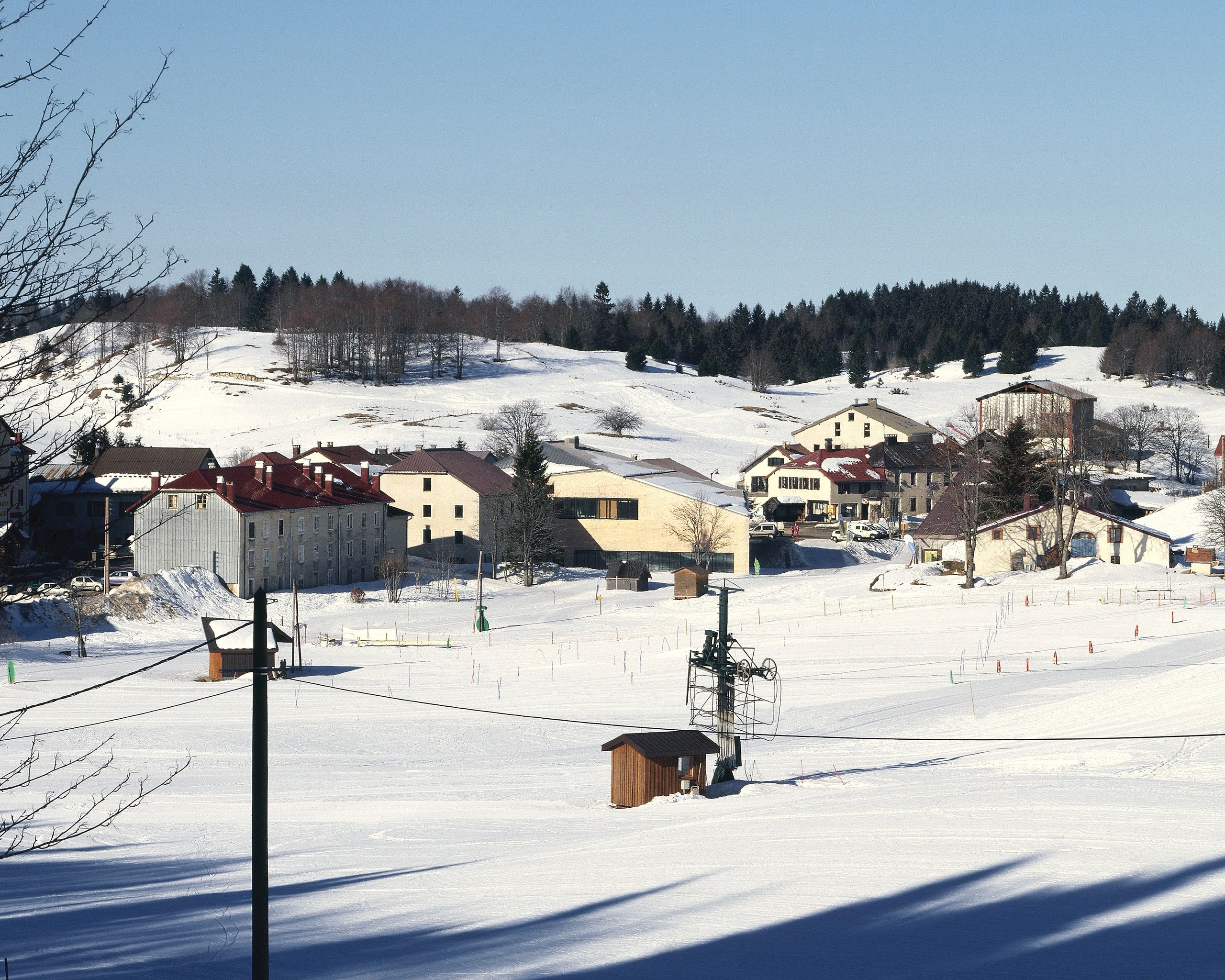 Vue lointaine de la Maison du Parc du Haut-Jura à Lajoux, volume compact en bois clair inséré dans le village enneigé, en dialogue avec les toitures traditionnelles.