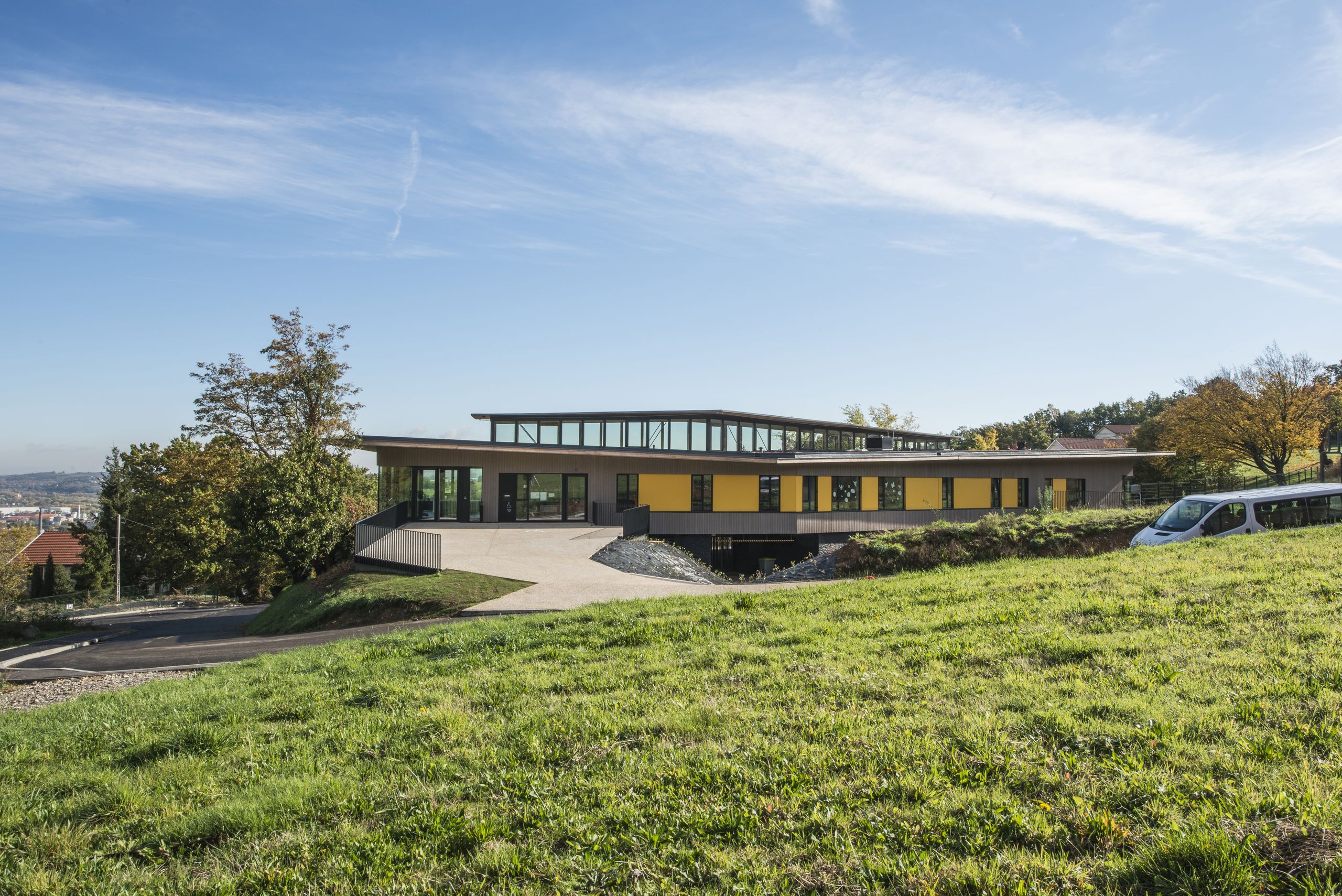 Centre de loisirs de Givors en belvédère : volume en équerre béton/ossature bois, façades rythmées de panneaux jaunes et bandeau vitré continu sur la toiture.