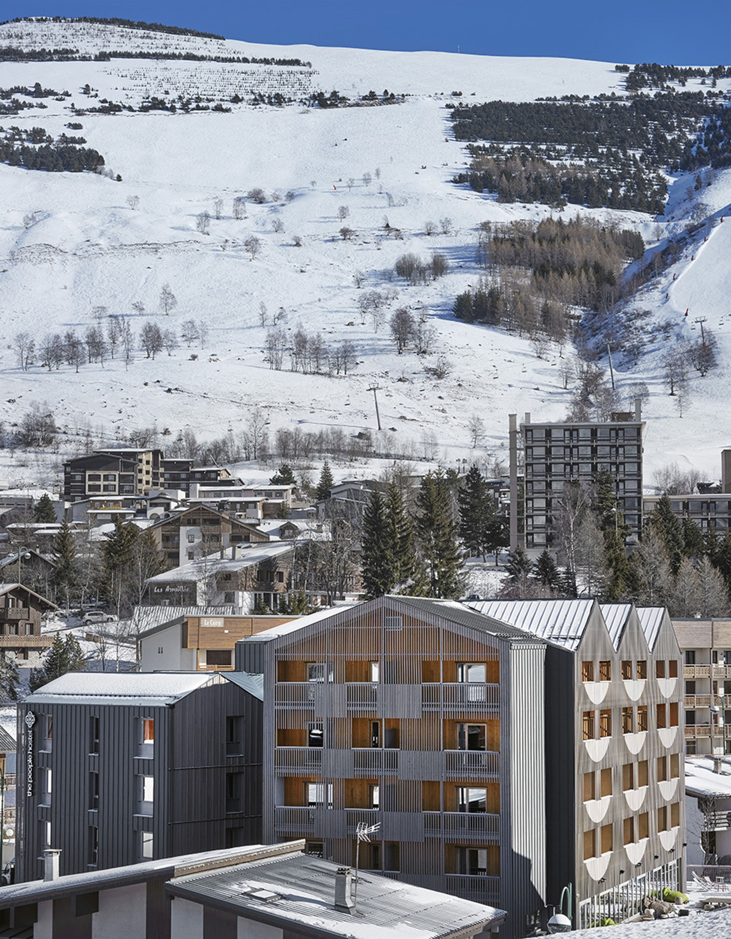 Auberge de jeunesse réhabilitée aux Deux Alpes, volumes fragmentés en CLT, double peau bois à claire-voie et bardage métallique face au paysage de montagne.