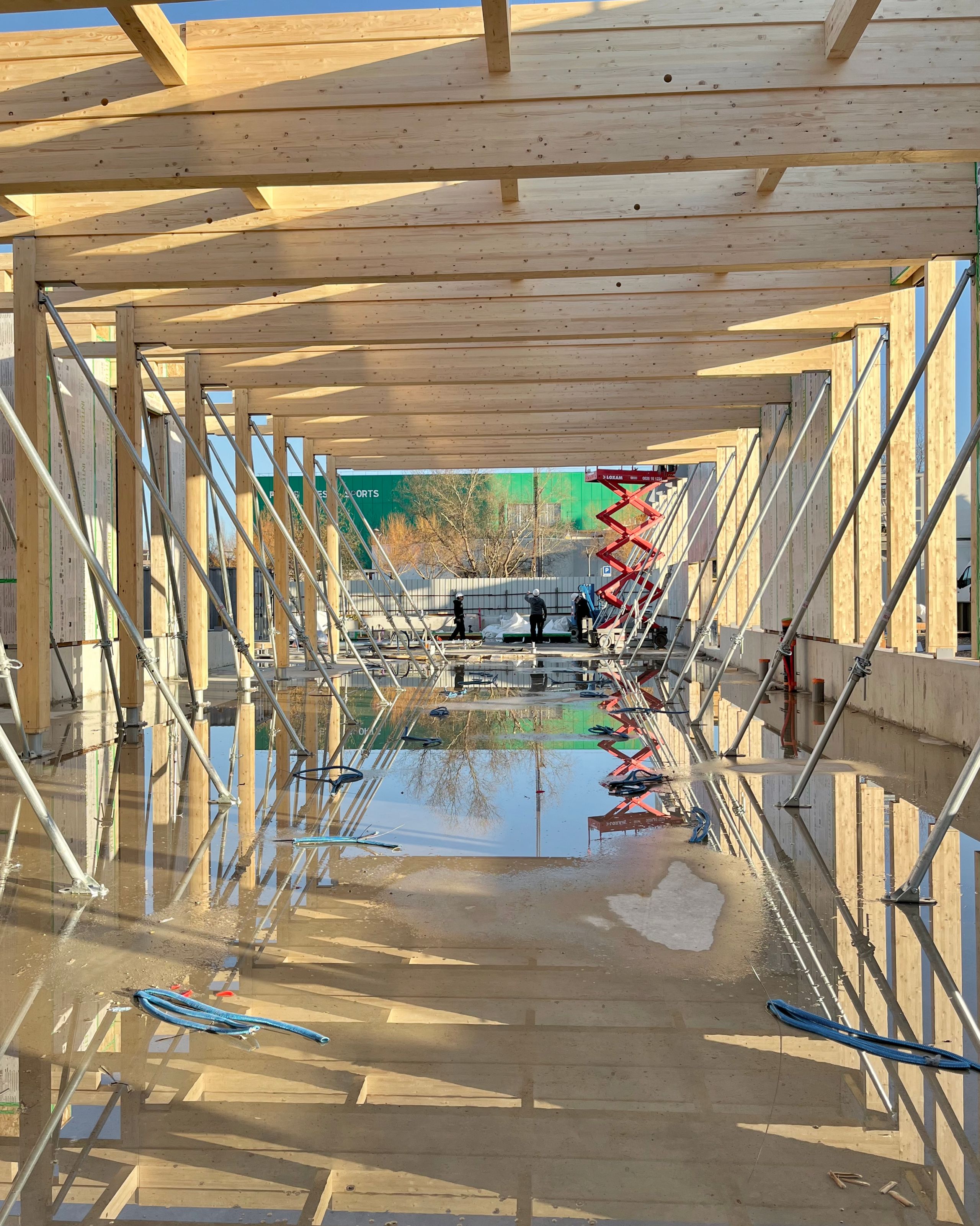 Chantier de la Cité campus Assia Djebar à Vaulx-en-Velin : structure bois en trame carrée, poutres apparentes et reflets au sol dans un volume traversant.