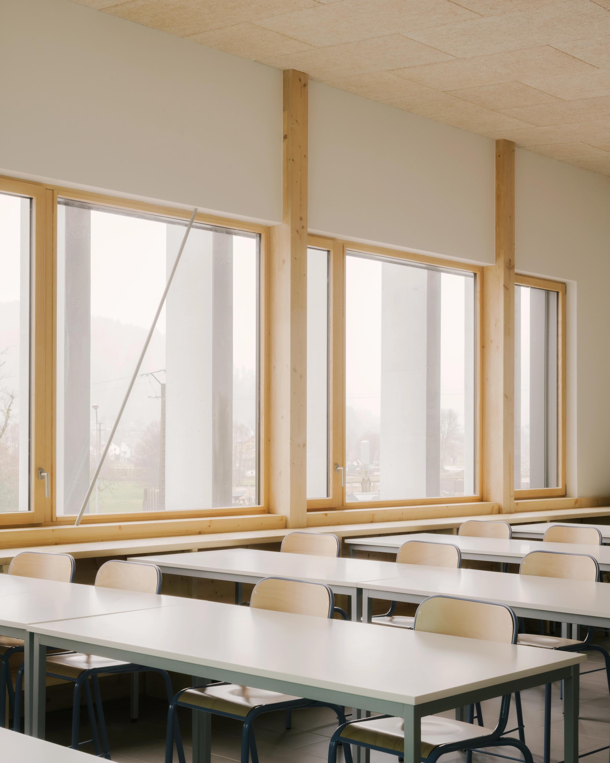 Salle de classe du Collège du Ban à Vagney, baignée de lumière naturelle, avec menuiseries bois, structure apparente et mobilier sobre aligné face aux baies.