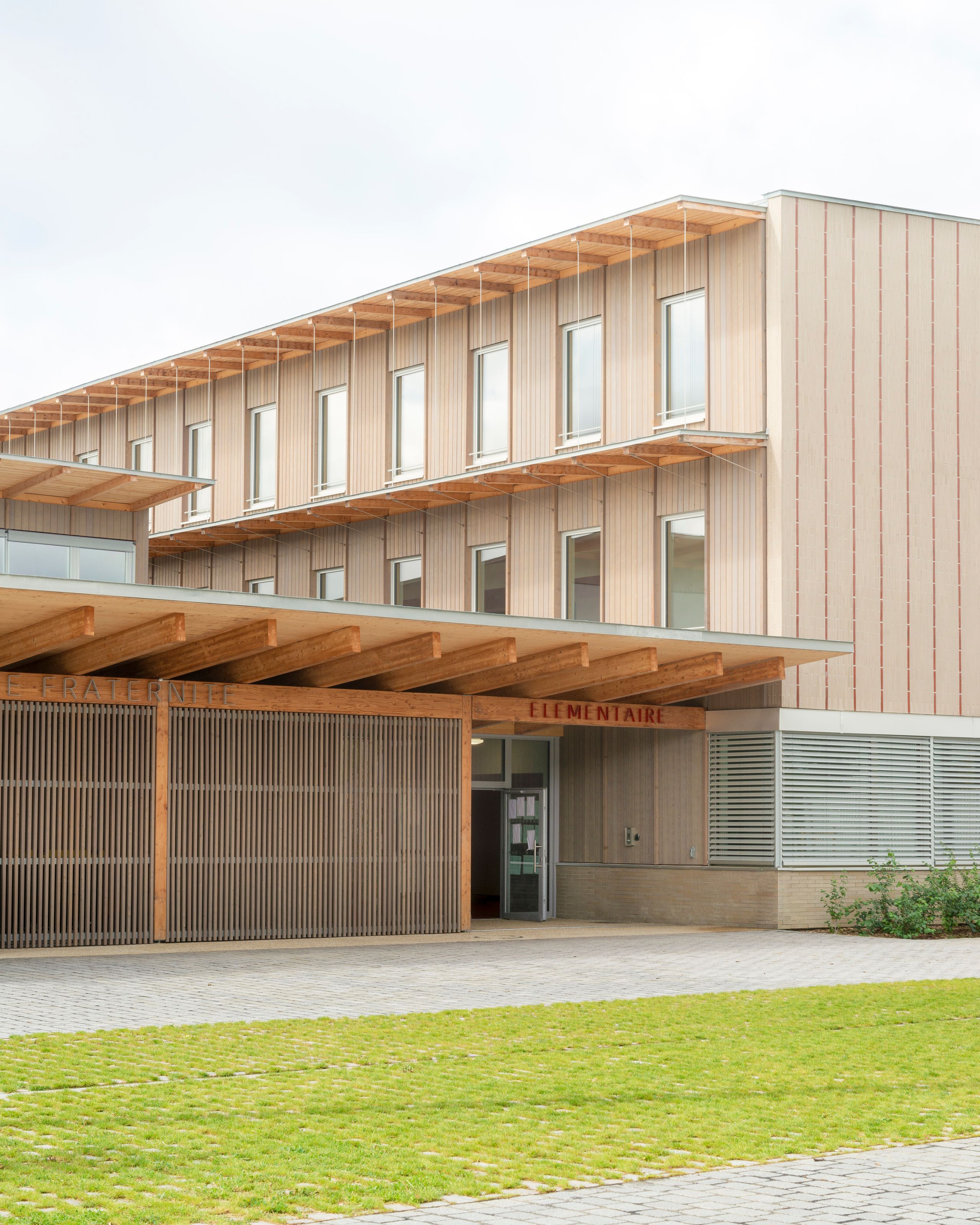 Façade bois et terre cuite du School complex Jacqueline Auriol au Bourget, avec préau en ossature bois, brise-soleil verticaux et large auvent d’entrée.