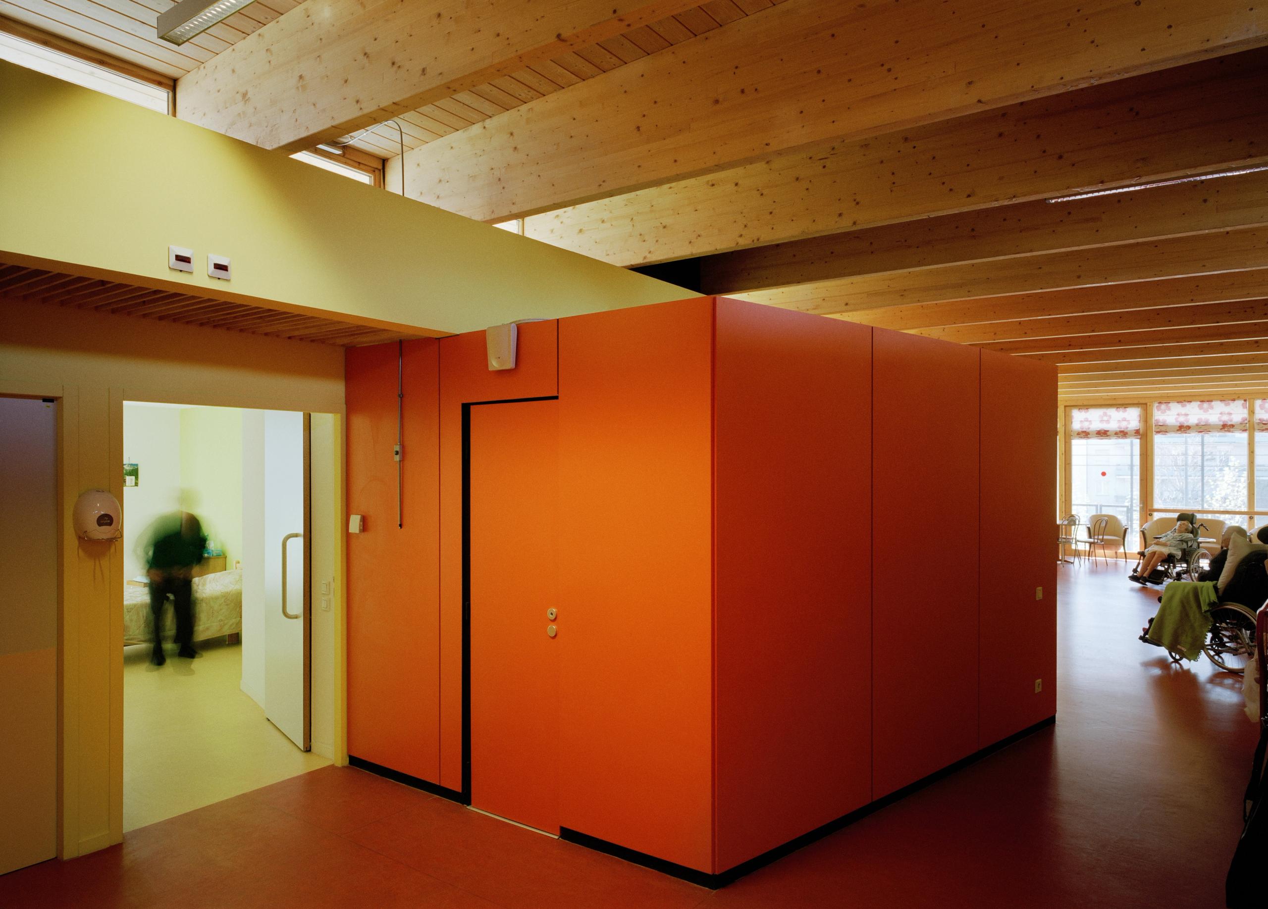 Intérieur de l’EHPAD à Grenoble par Tectoniques : volume central orange, structure bois apparente, circulation éclairée naturellement vers salle commune vitrée.