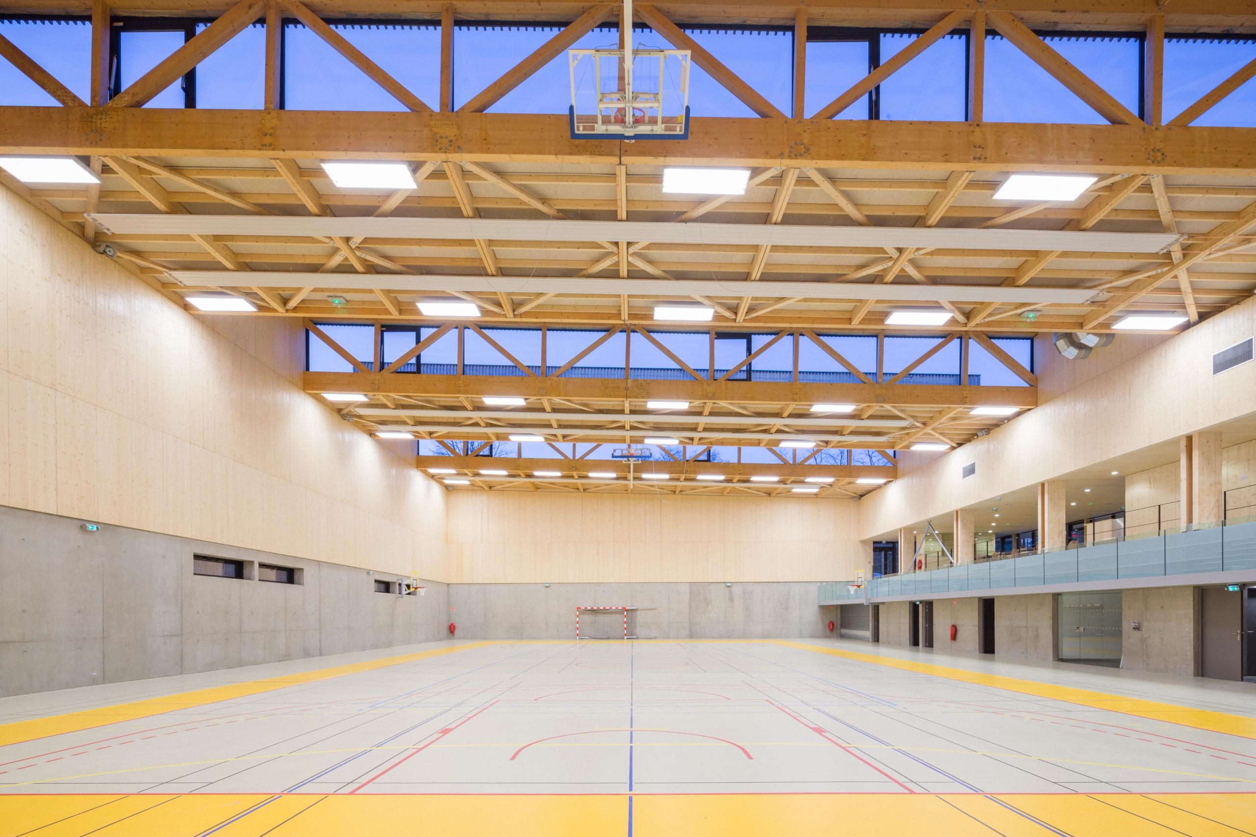 Intérieur du gymnase Hacine Cherifi à Rillieux-la-Pape, grande halle sportive en béton et bois, charpente treillis apparente et bandeau vitré nord continu.