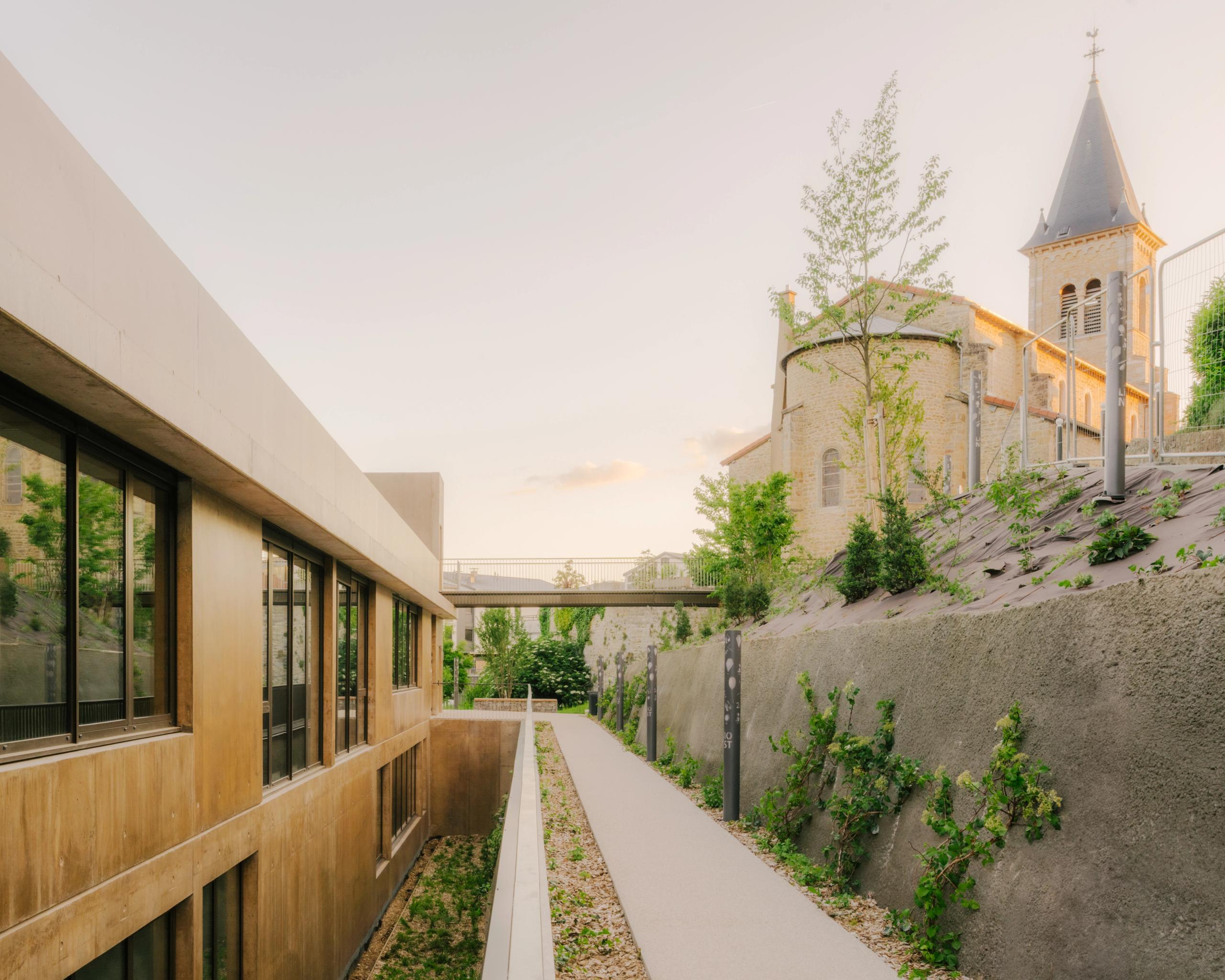 Maison des familles à Limonest : volumes en béton teinté en gradins sous le parvis de l’église, cheminement en terrasse et talus planté sur forte pente.