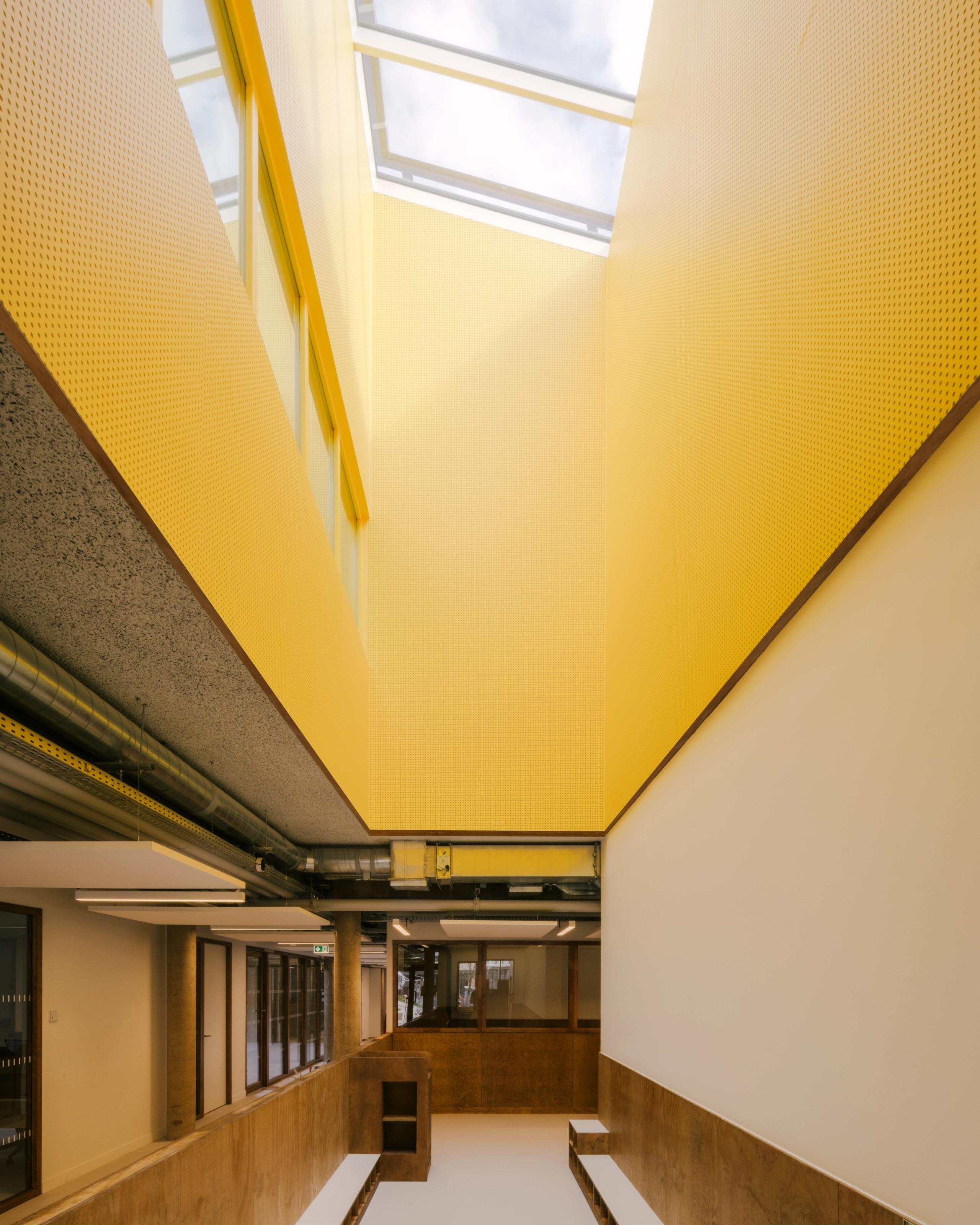 Atrium de la Maison des familles à Limonest, double hauteur en béton et bois, parois jaunes perforées et puits de lumière zénithal structurant les circulations.
