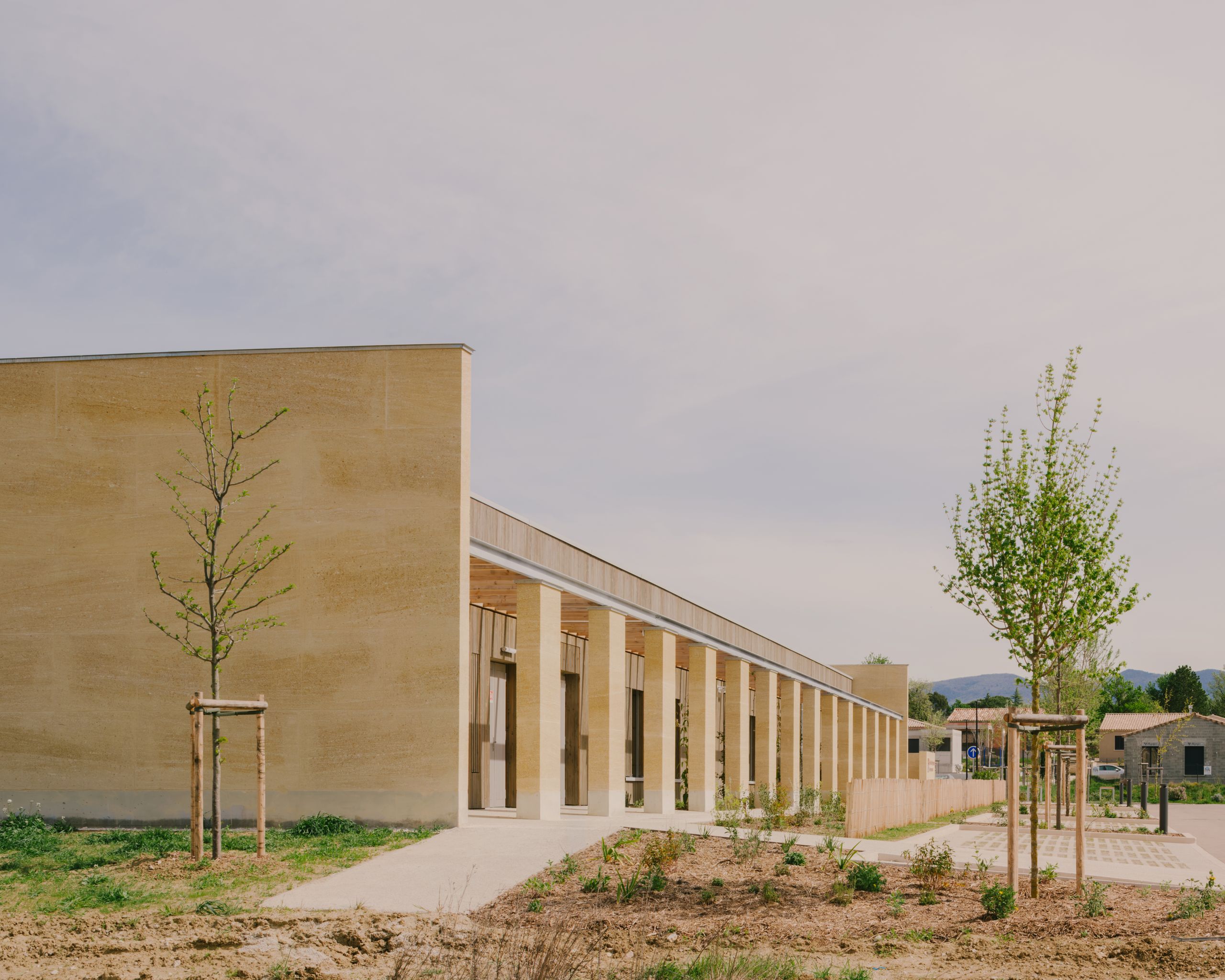 Façade du Pôle culture et jeunesse à Vaison-la-Romaine : volume bas en pierre massive, portique rythmé de poteaux, jardin et jeunes arbres en premier plan.
