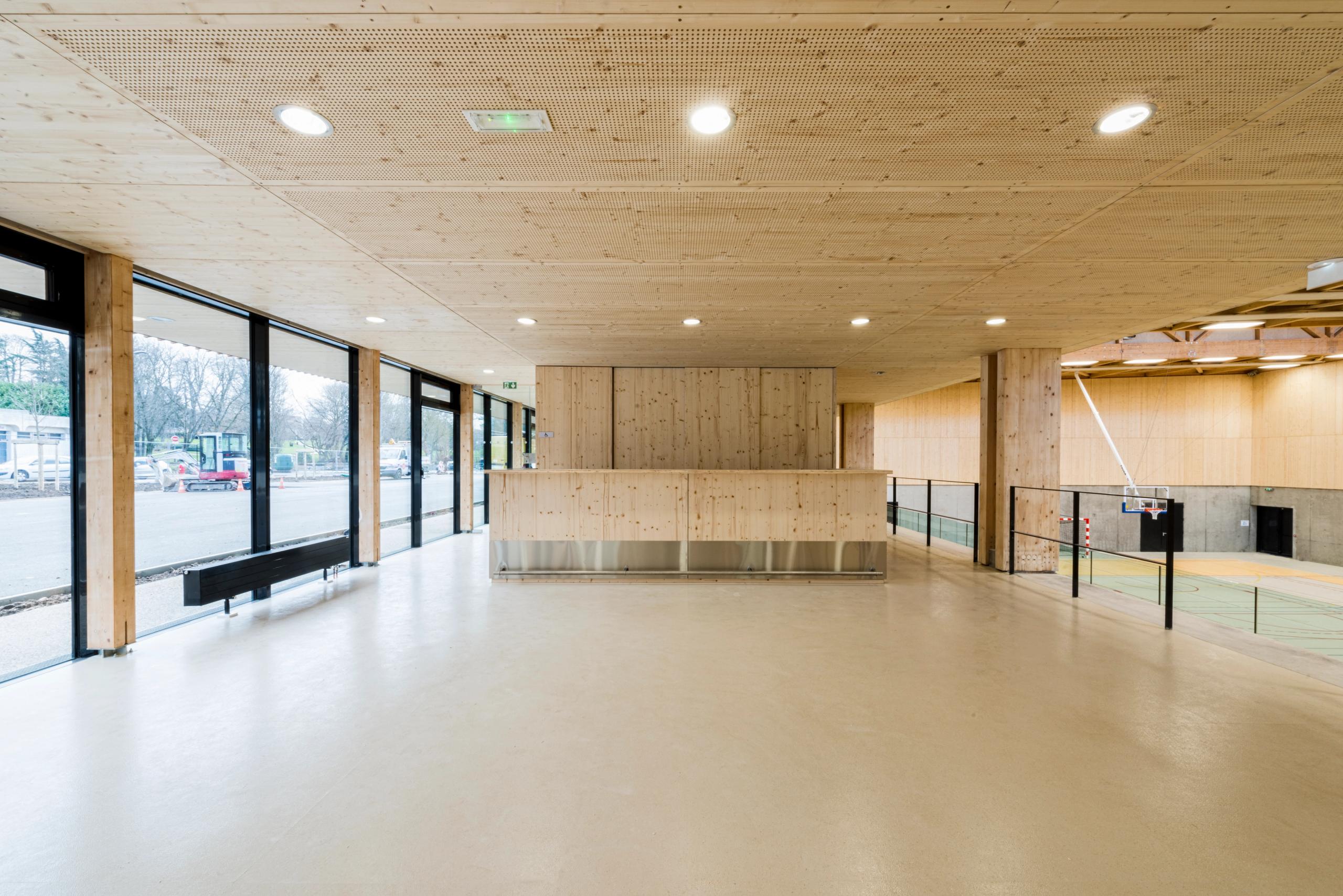 Hall du gymnase Hacine Cherifi à Rillieux-la-Pape, vaste espace en structure bois apparente, ouvert par une façade vitrée sur rue et sur la salle de sport.