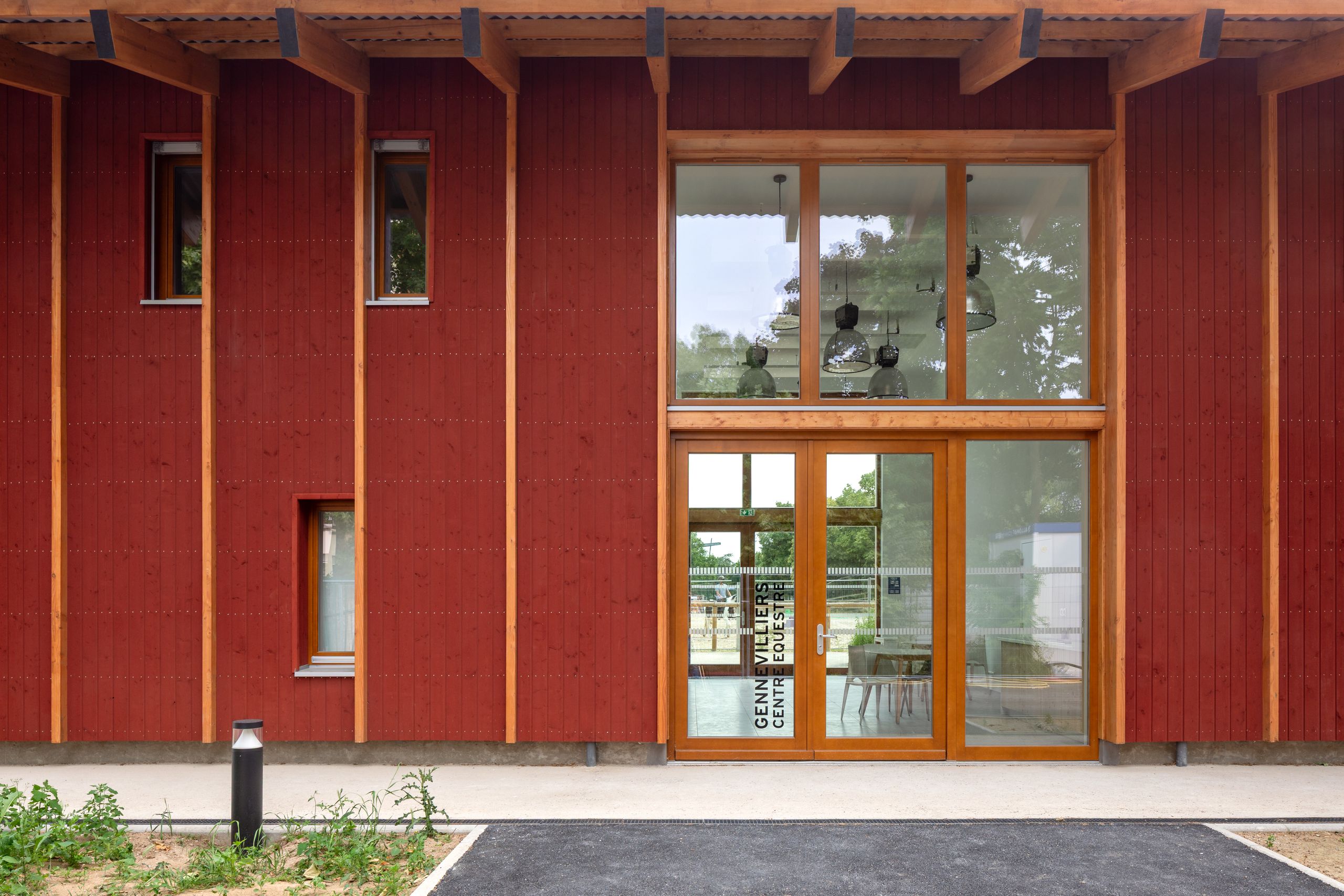 Façade du centre équestre de Gennevilliers : volume en bois bardé de rouge de Falun, trame verticale marquée et large baie vitrée du club house sur cour urbaine.