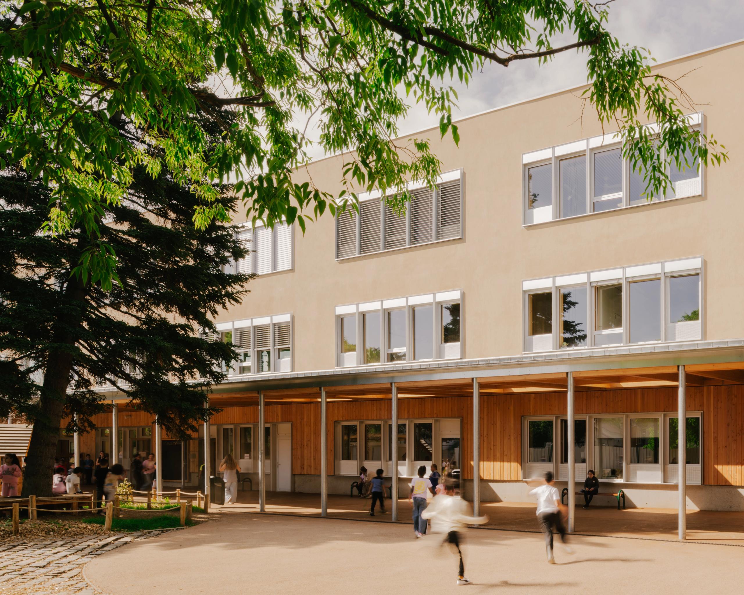 Cour du groupe scolaire Kennedy à Lyon, façade bois et enduit minéral à R+2, préau poteaux-poutres ouvert sur une cour de récréation végétalisée.