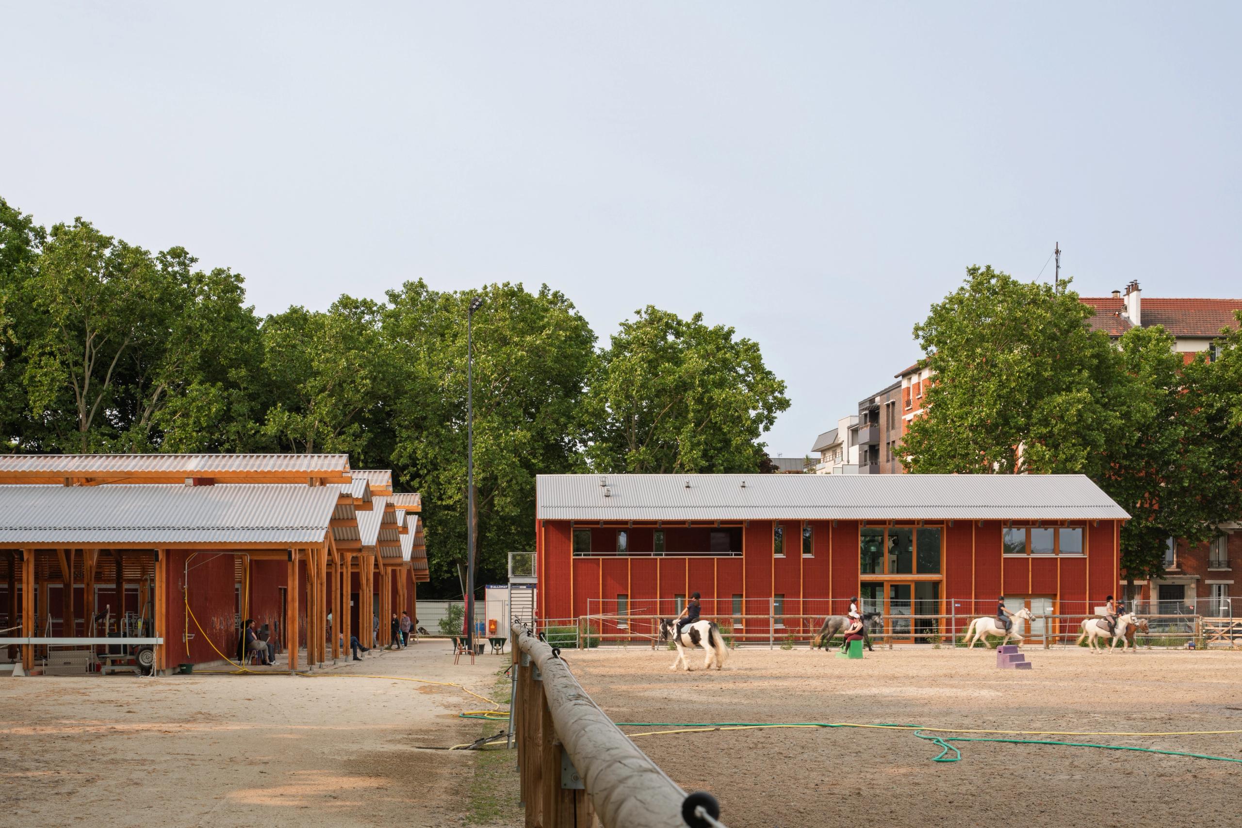 Falun red timber stables and arena of the Gennevilliers equestrian center, with corrugated roofs framing an outdoor riding ring in a dense urban setting.