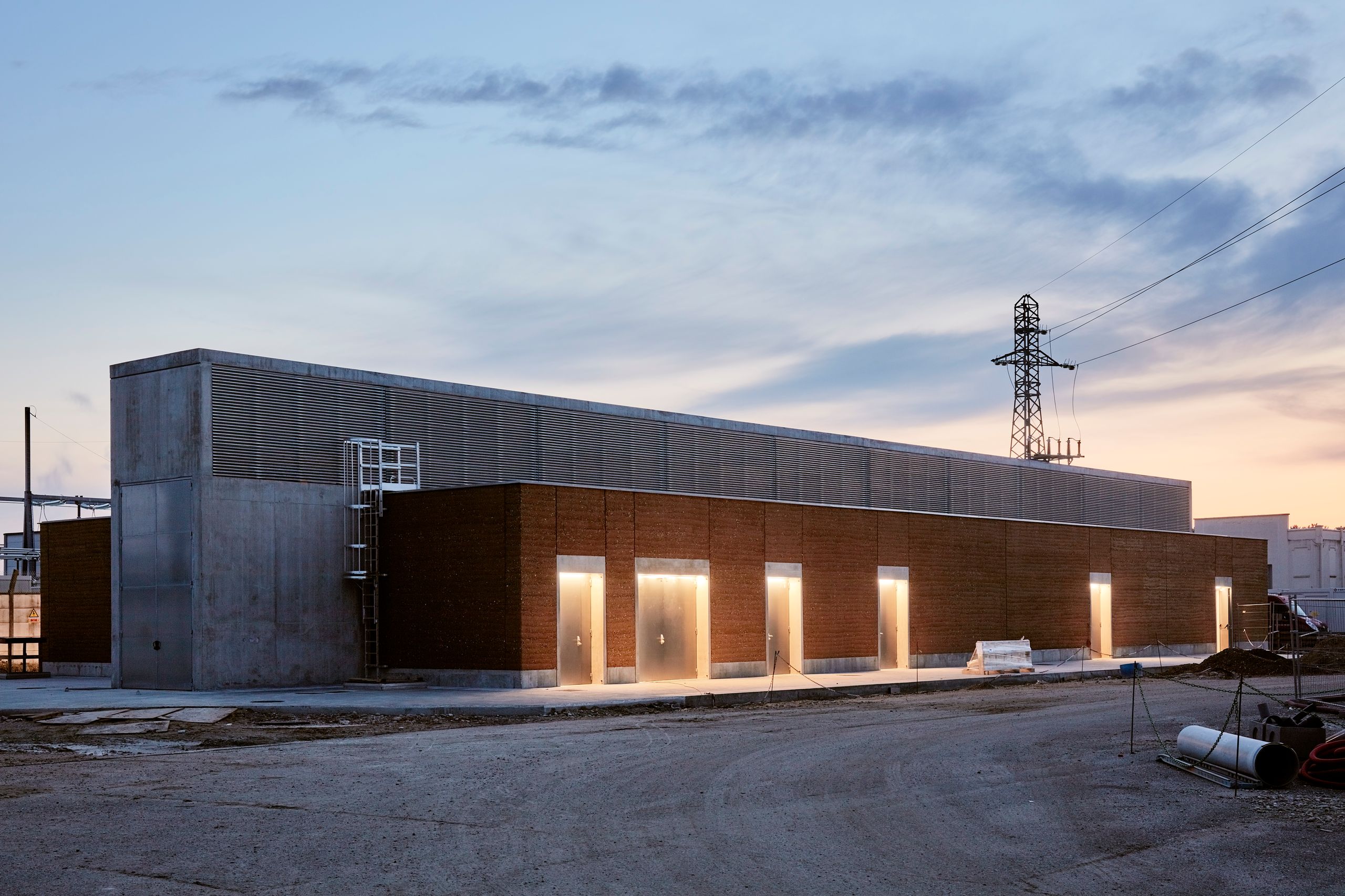 Poste source de Cusset à Villeurbanne, volume linéaire en béton brut, pisé et acier galvanisé, façades rythmées par des portes métalliques éclairées au crépuscule.